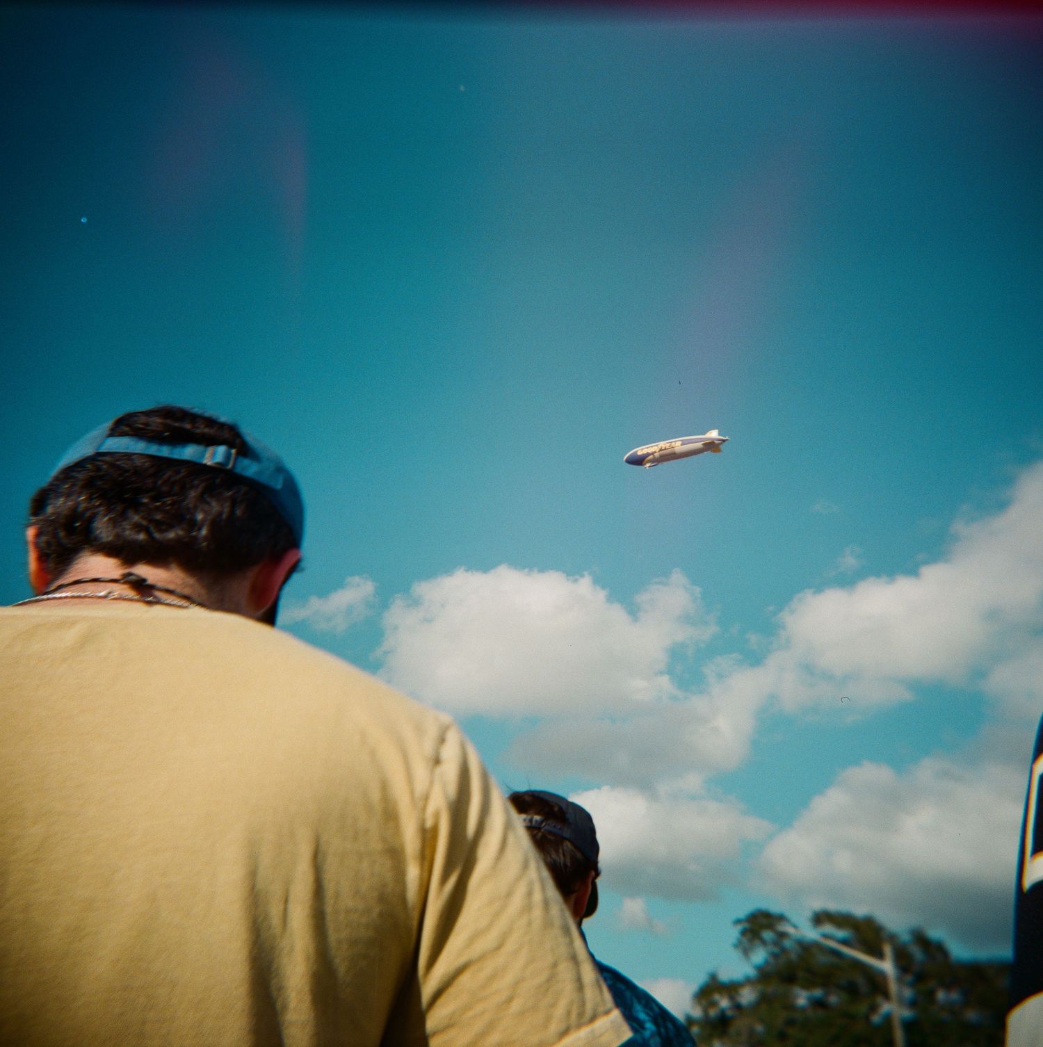 Two people watch a blimp flying overhead against a bright blue sky with scattered clouds.