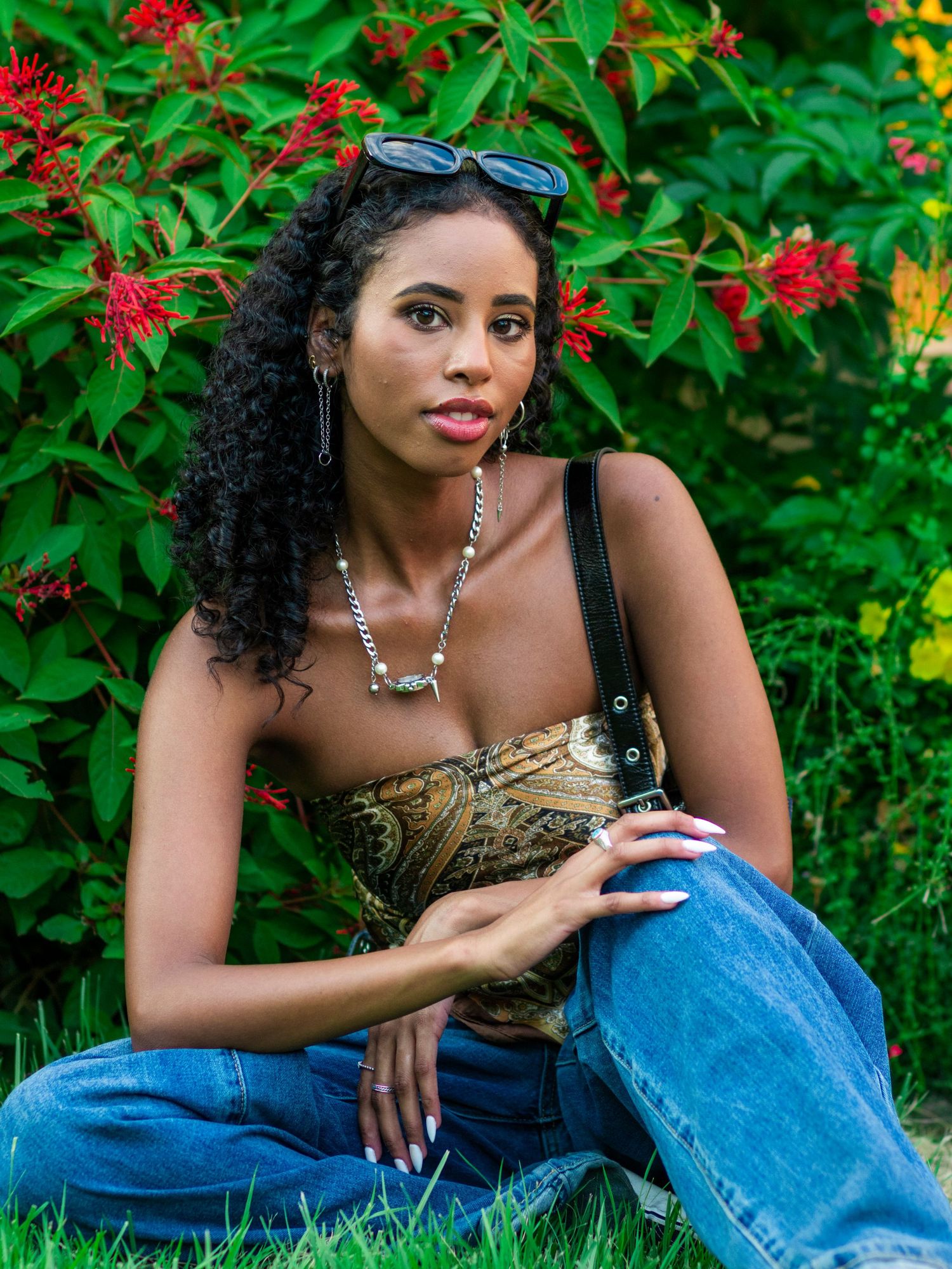 Young Black woman with curly hair in patterned corset top and jeans sitting on grass outdoors.