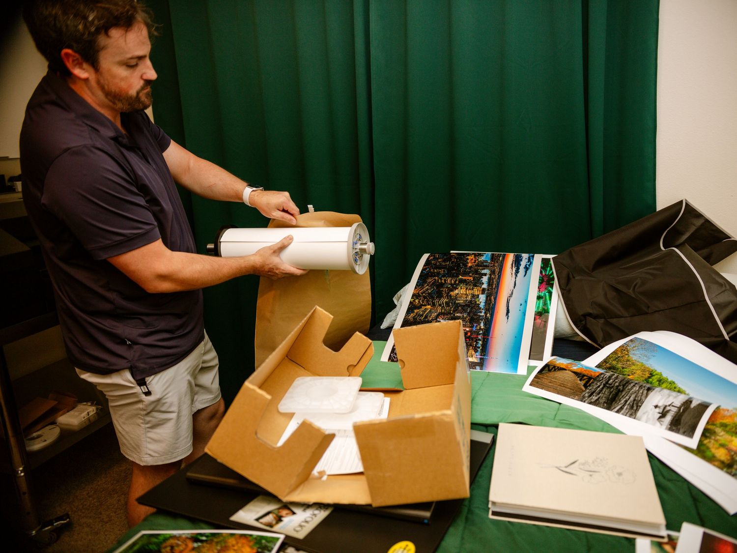 Man in navy polo unpacking large prints from a cardboard box on a green-draped table.