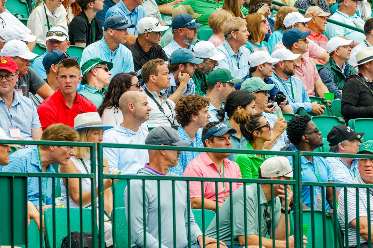 Crowd of spectators watching a golf tournament from the stands, wearing casual summer attire at a green venue.
