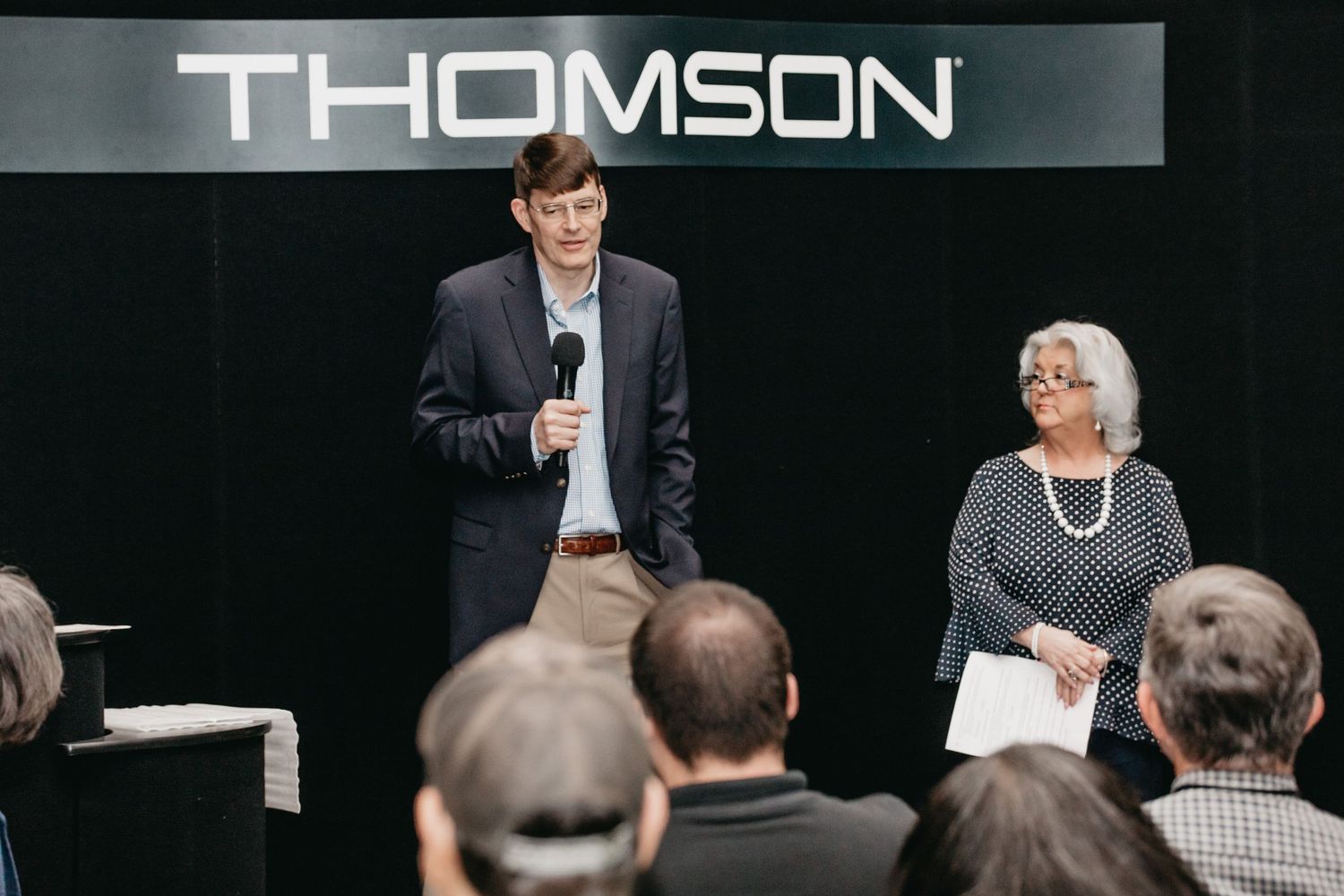 Two speakers present at a Thomson corporate event, man with microphone in suit, woman beside him holding papers.