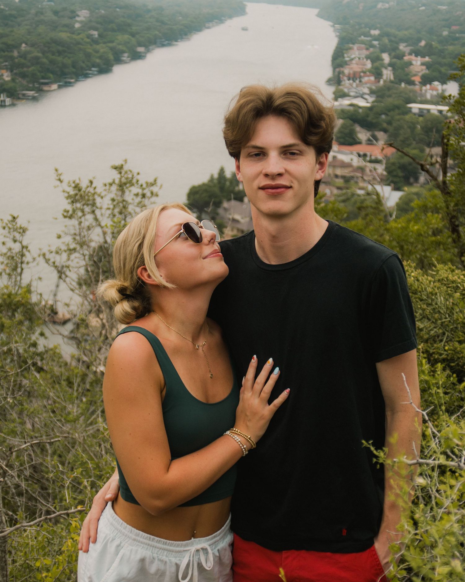 A young couple poses on a scenic overlook with a river and lush greenery visible in the background.