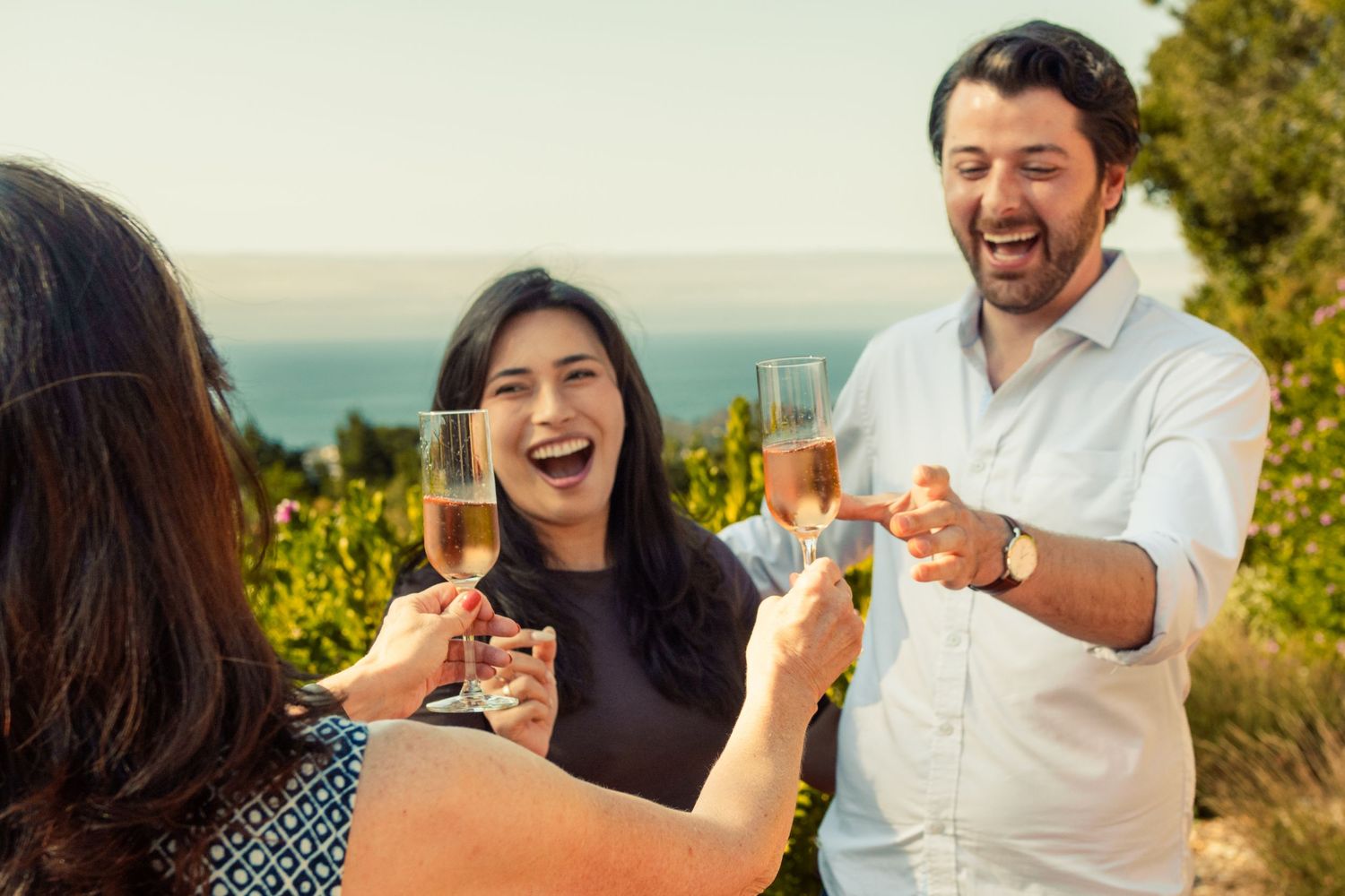 Three friends laughing and toasting with rosé wine glasses outdoors at a vineyard with ocean views.