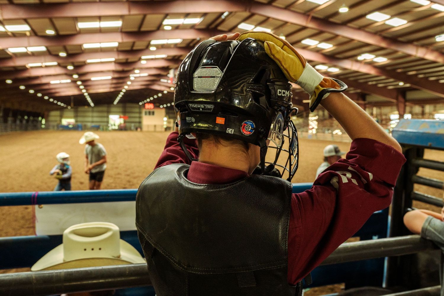 Young rodeo rider adjusting protective helmet in indoor arena, preparing for bull riding competition.