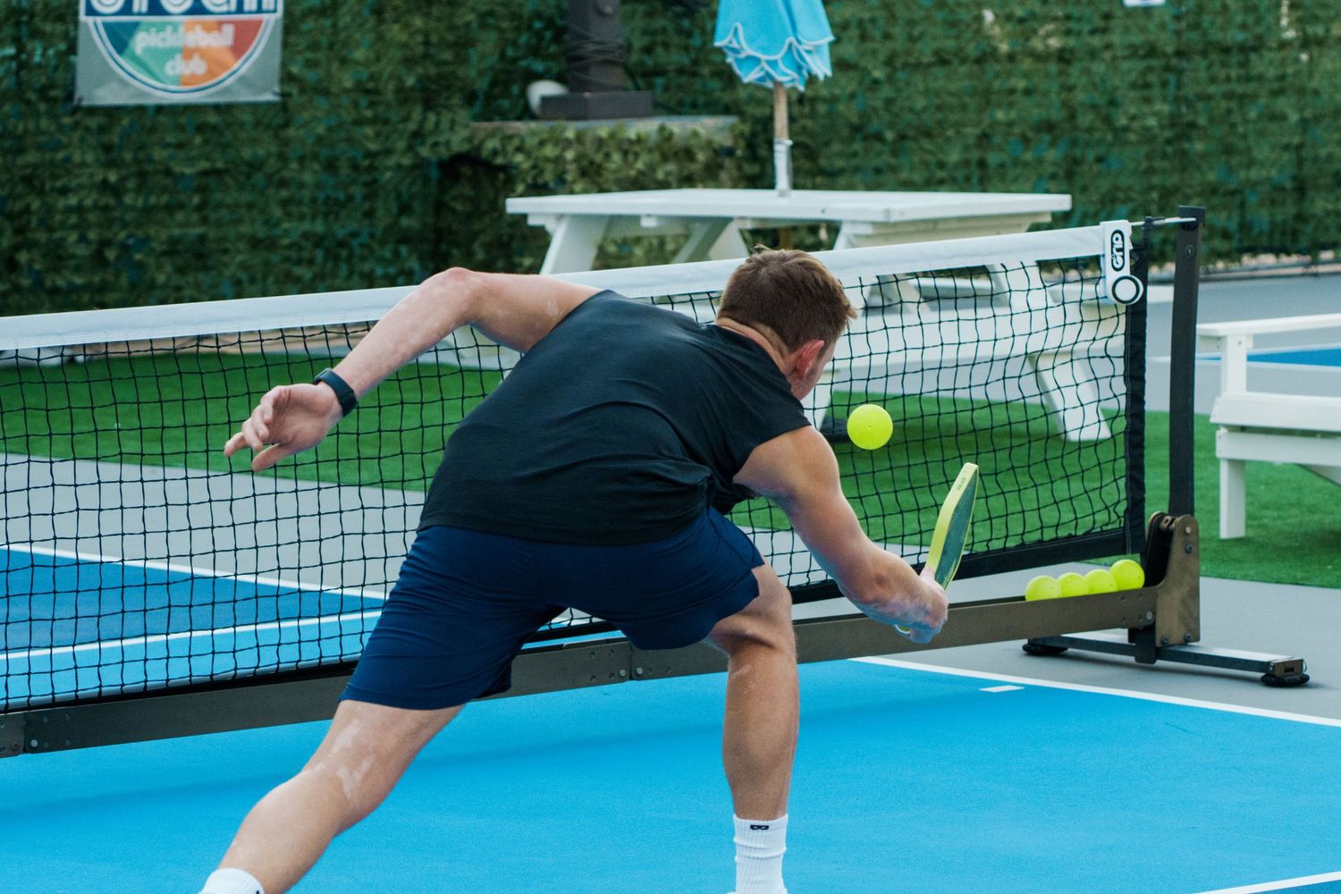 Man playing pickleball on a blue court, reaching to hit a yellow ball near the net outdoors.
