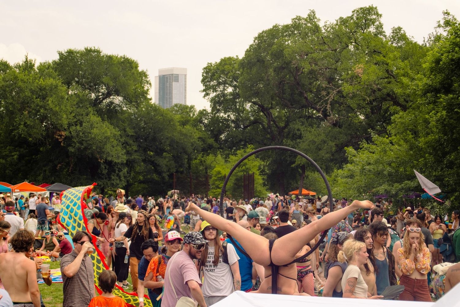 Crowd of festival-goers in a park with a performer doing a split, trees and city skyline in background.