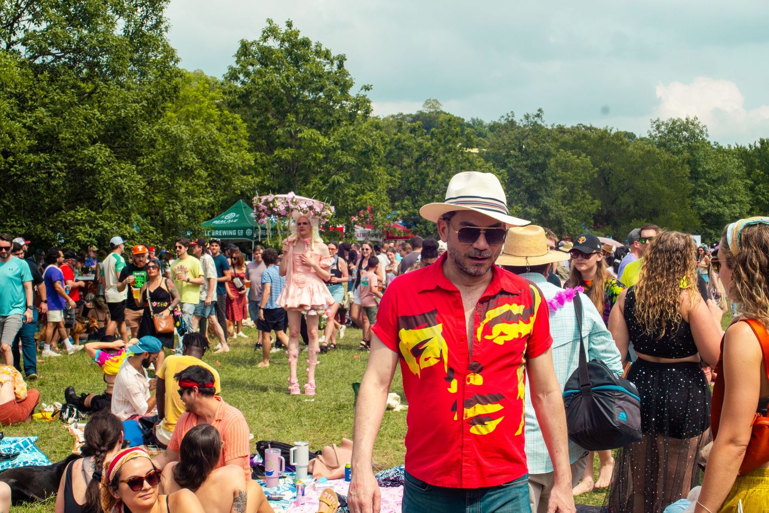 Man in red shirt and straw hat at a crowded outdoor music festival surrounded by colorful attendees on a sunny day.