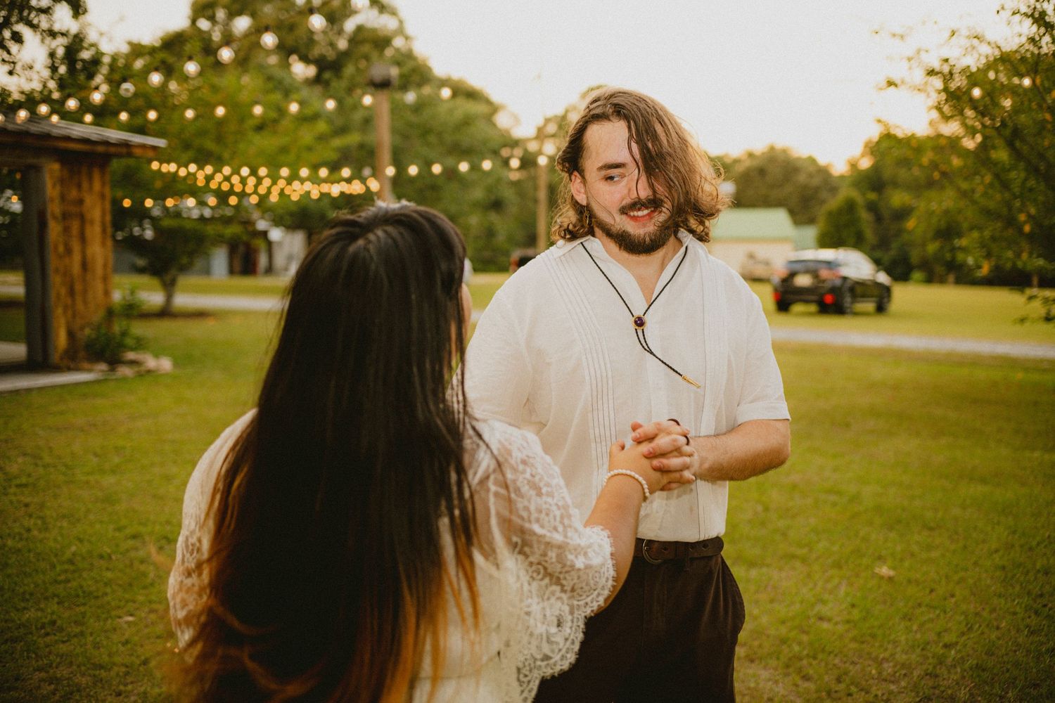 Smiling groom holds bride's hands while dancing outdoors at golden hour wedding reception.