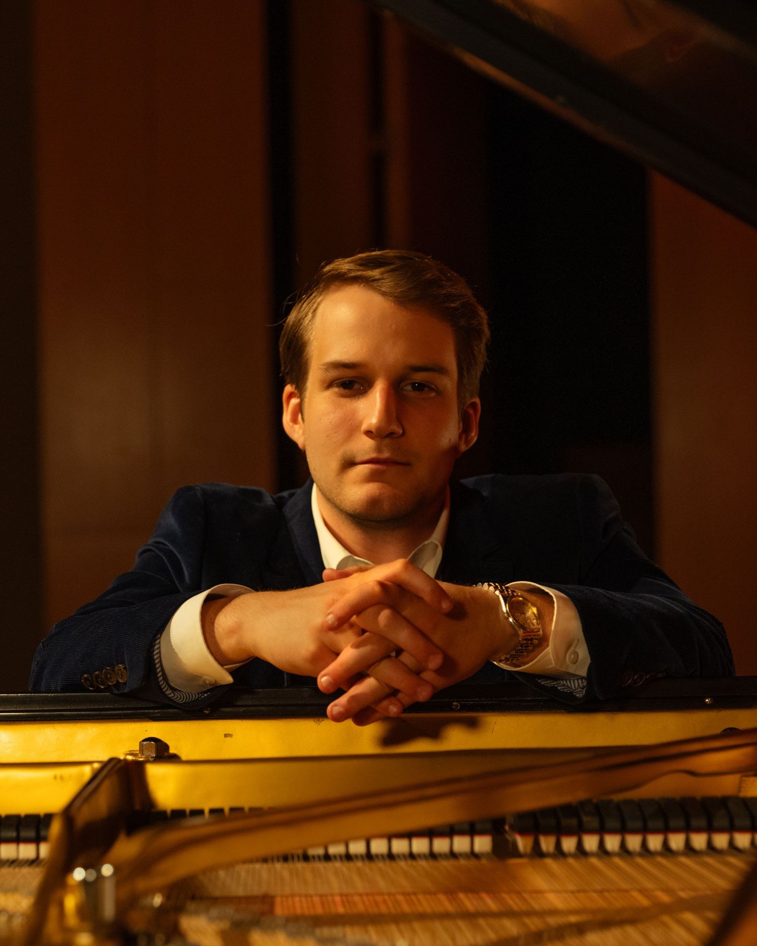 Young male pianist resting chin on hands at a grand piano in warm studio lighting.