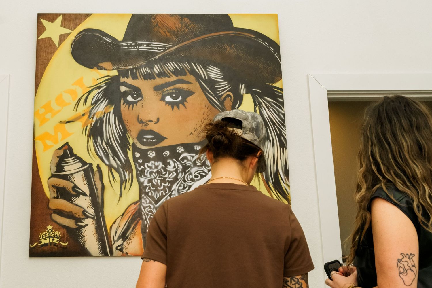 Gallery visitor examines bold pop art painting of cowboy woman with spray can at street art exhibition.