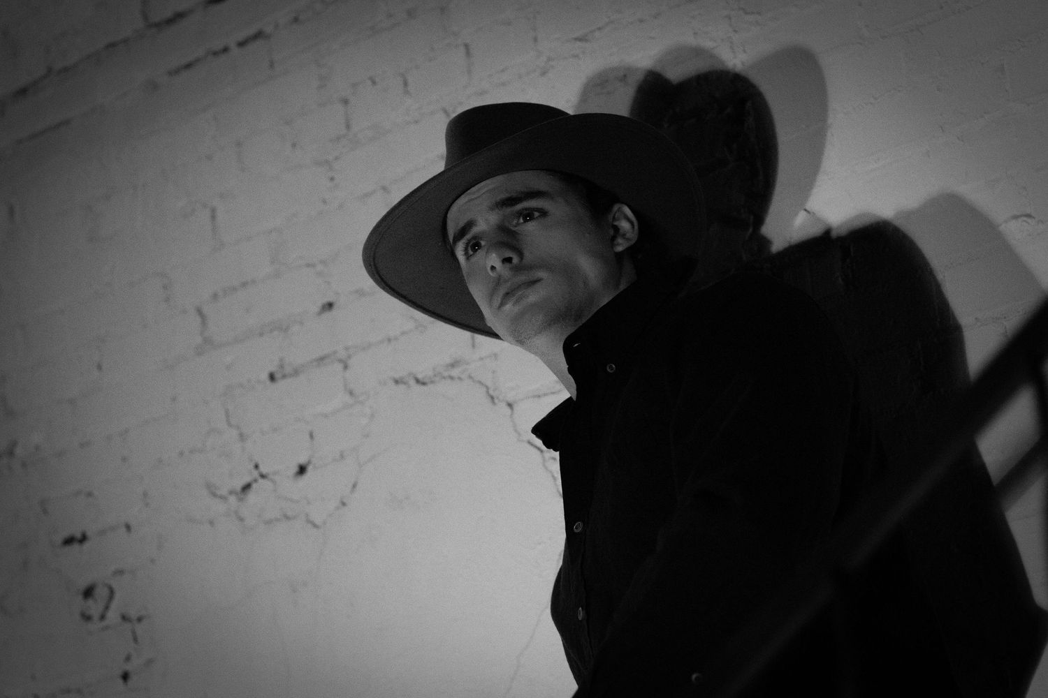 Black and white portrait of a man in a wide-brim hat and dark coat leaning against a white brick wall.