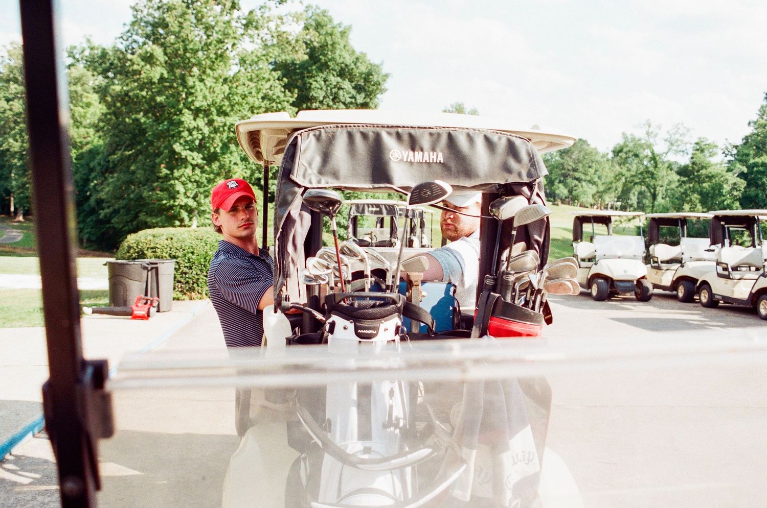Two golfers loading equipment onto a golf cart at a sunny golf course with trees in the background.