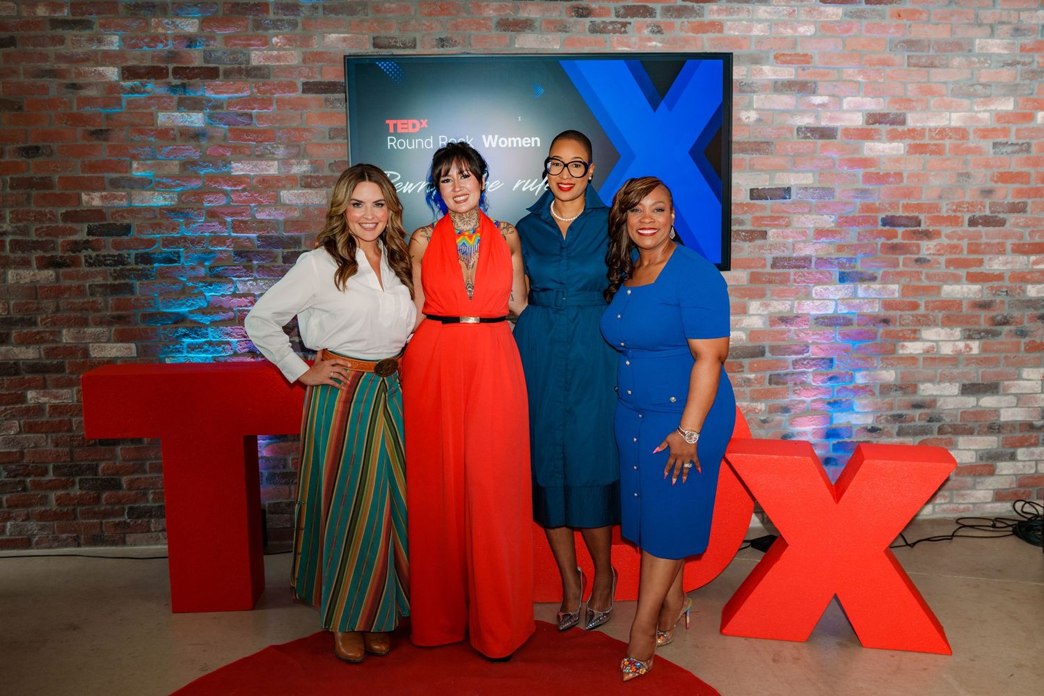Four women posing together at a TEDx event in front of a branded backdrop with red letter displays.