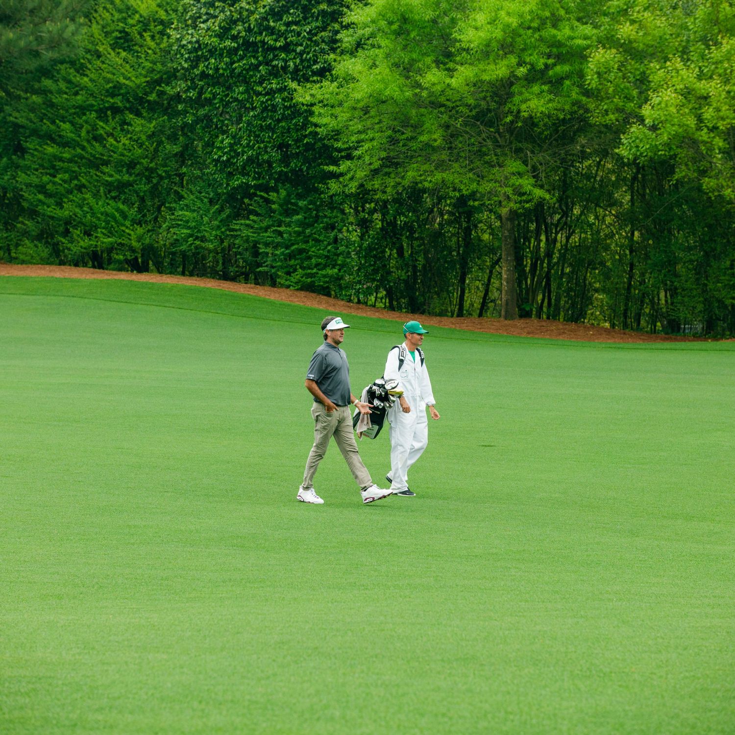 A golfer and caddie walk along a lush green fairway surrounded by tall trees at a golf course.