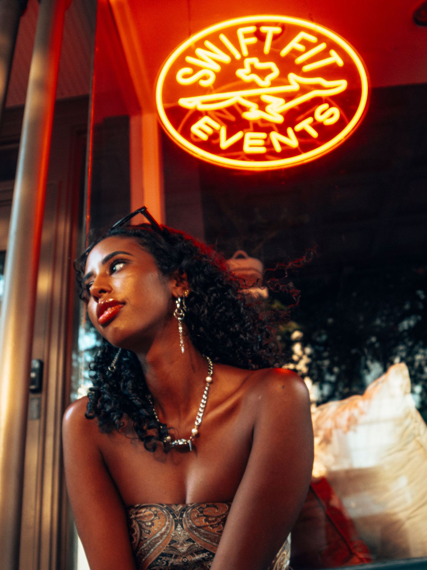 Young Black woman with curly hair gazes upward beneath a glowing red Swift Fit Events neon sign at night.