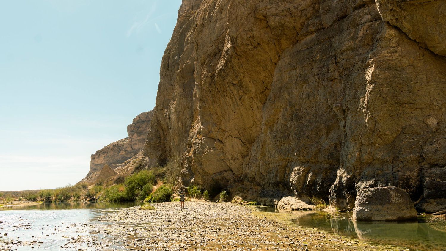 Scenic view of a towering limestone cliff beside a calm river with hikers exploring the shoreline.