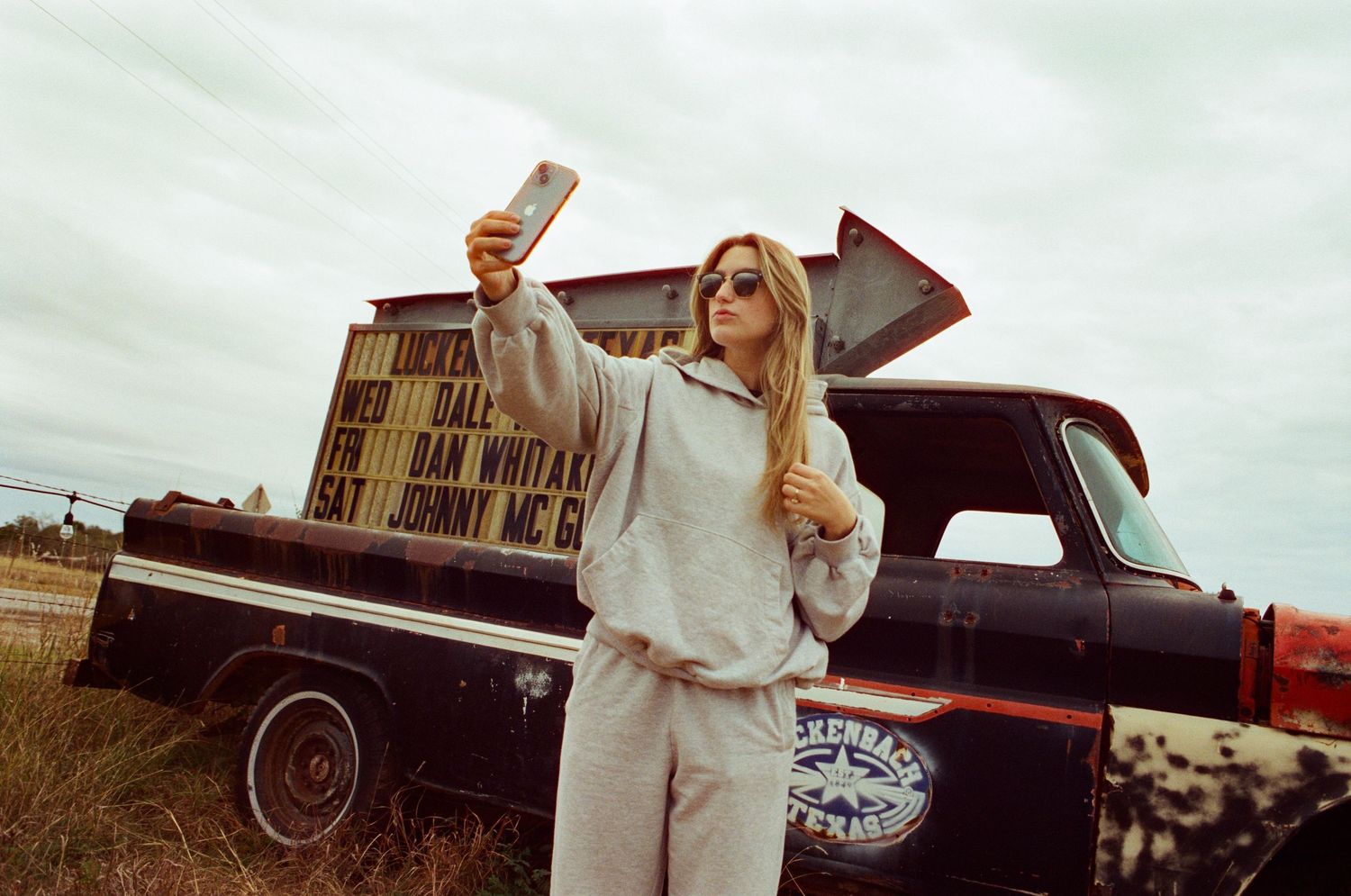 Young blonde woman in grey hoodie takes a selfie beside a vintage truck outdoors.