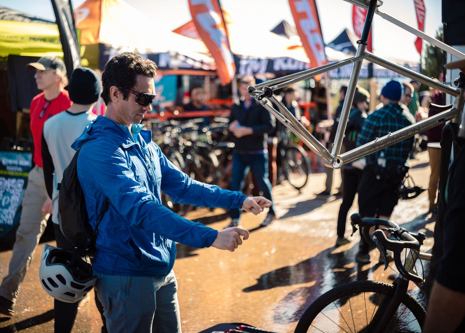Cyclist in blue jacket inspects a titanium bike frame at a sunny outdoor cycling expo event.