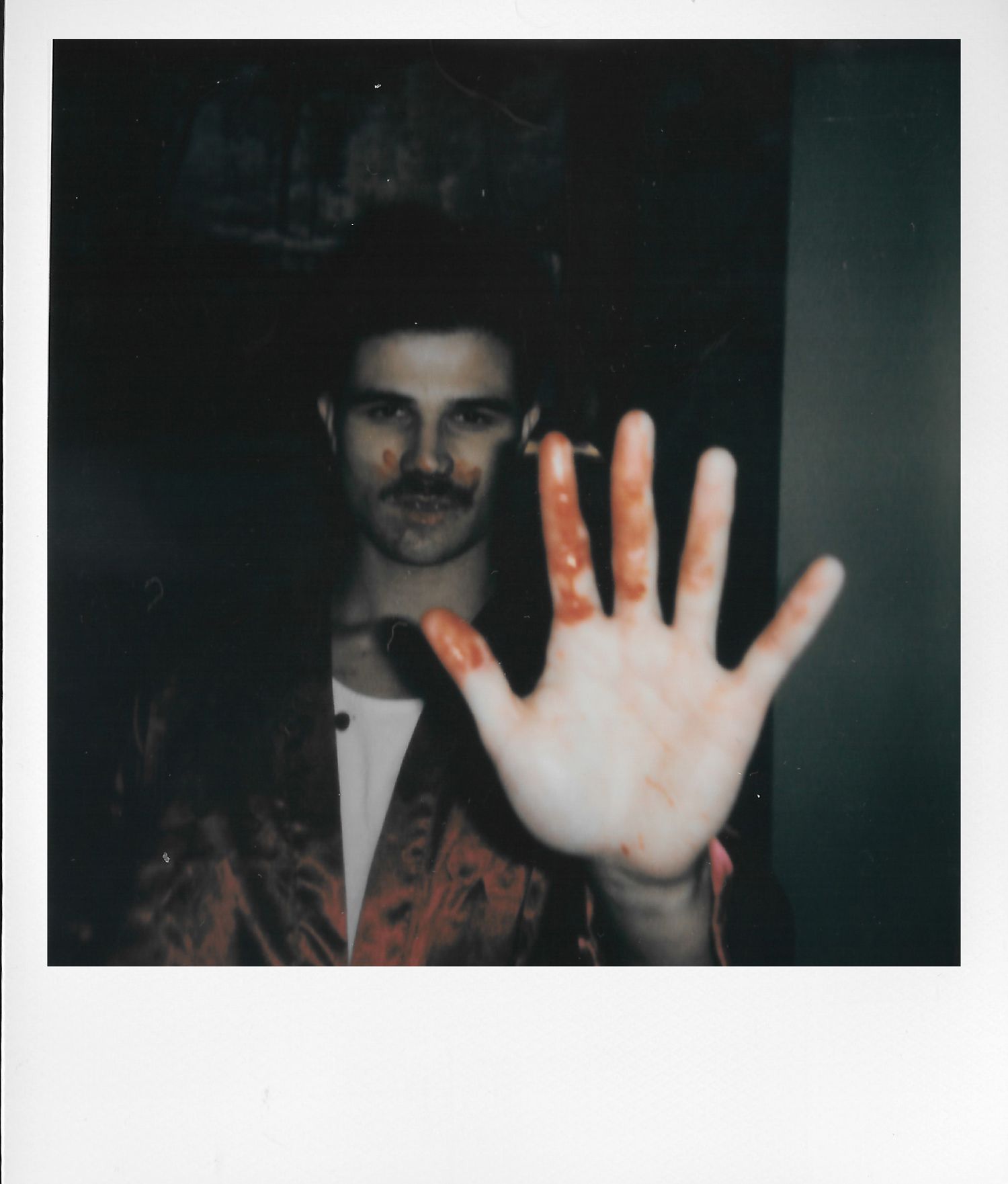 Man with mustache in patterned jacket holds up hand with red paint stains in vintage Polaroid photo.