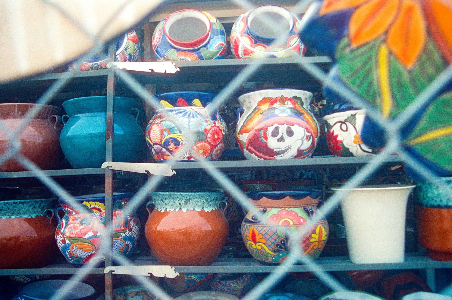 Colorful hand-painted Mexican pottery and ceramic bowls displayed on metal shelves behind a chain-link fence.