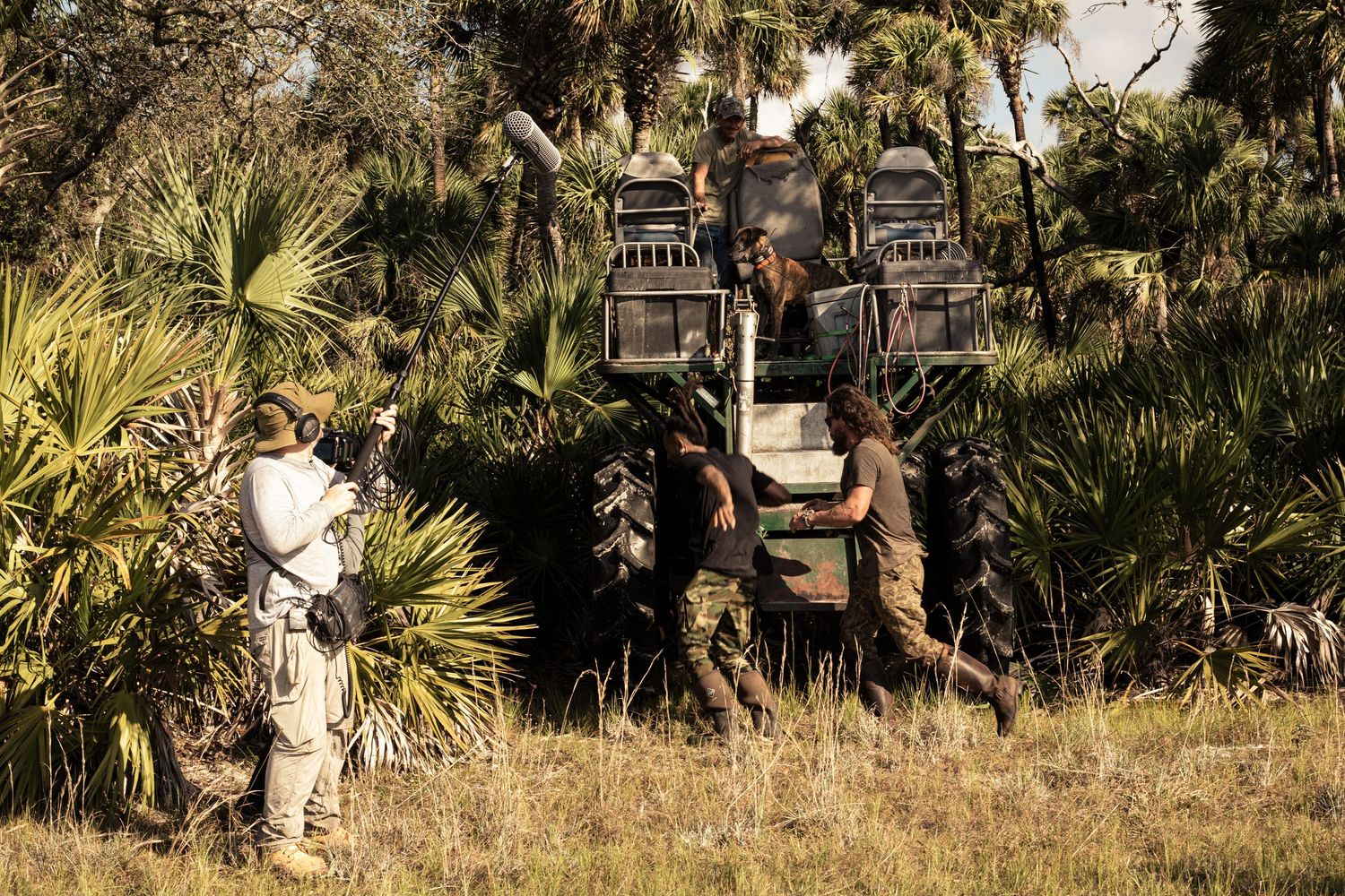 Two people work near a large tractor in a grassy field surrounded by palm trees in a natural outdoor setting.