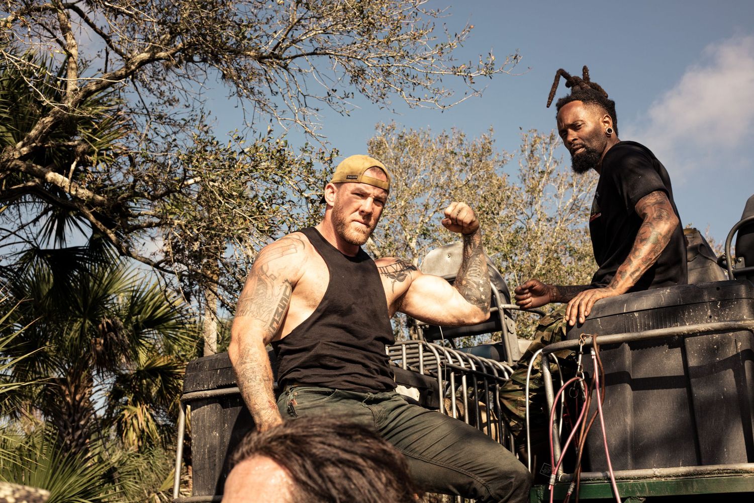 Two tattooed men flexing muscles outdoors near a vehicle, surrounded by trees on a sunny day.