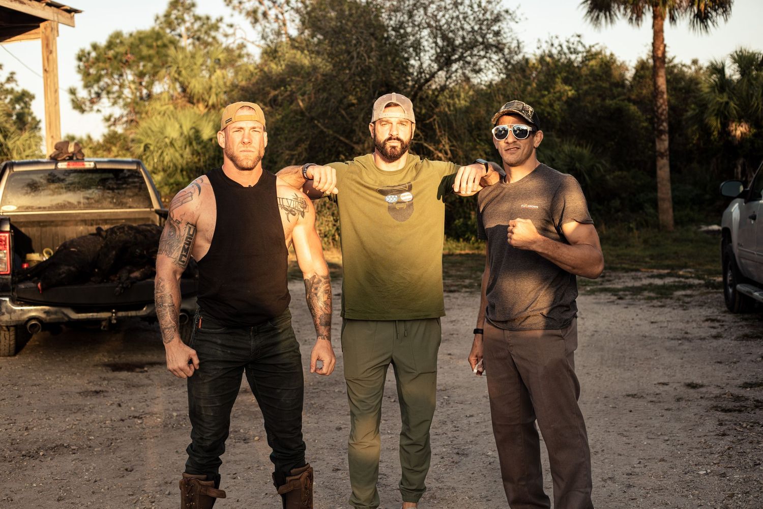 Three tattooed men posing together outdoors near a pickup truck in a rural setting during golden hour.