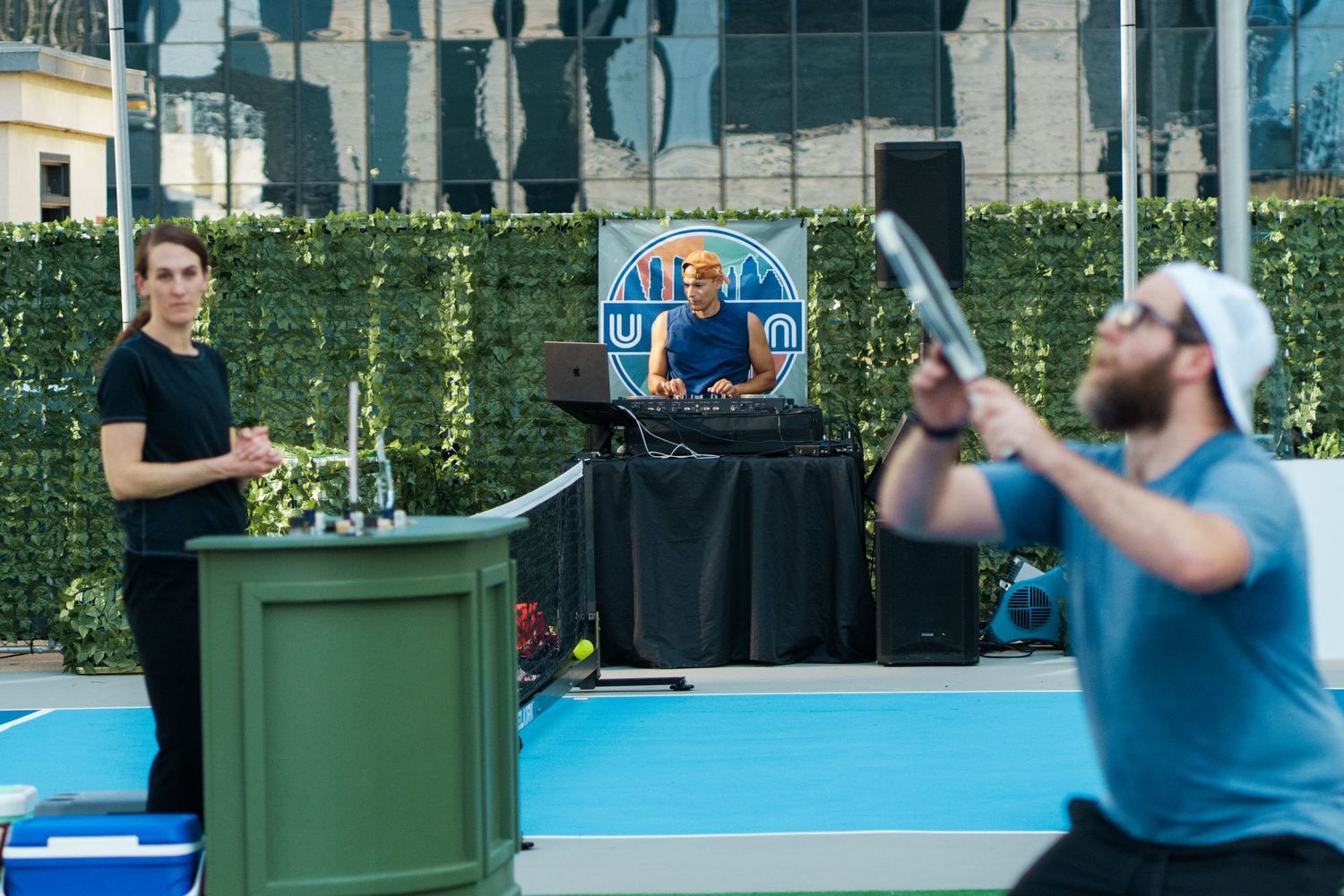 Two men on a blue tennis court with a DJ booth and green hedge backdrop at an outdoor urban event.