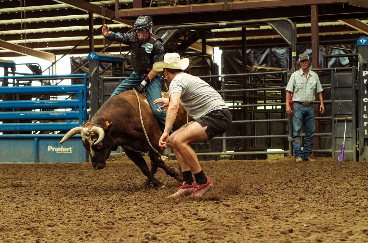 Bull rider competing in indoor rodeo arena while a rodeo clown assists with rope control during an intense bull riding event.