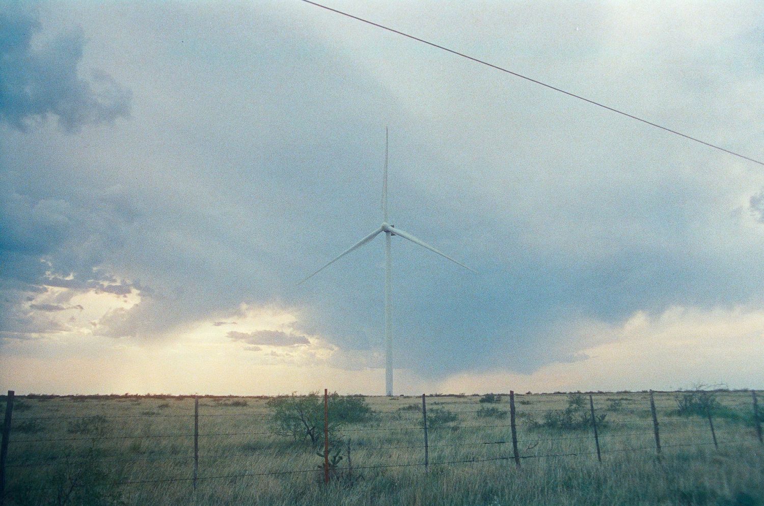 A lone wind turbine stands in a vast green field under a dramatic cloudy sky near a wire fence.