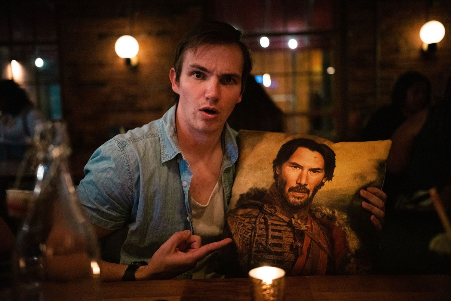 Young man at a restaurant table holding a decorative pillow featuring a portrait of a bearded man in ornate clothing.