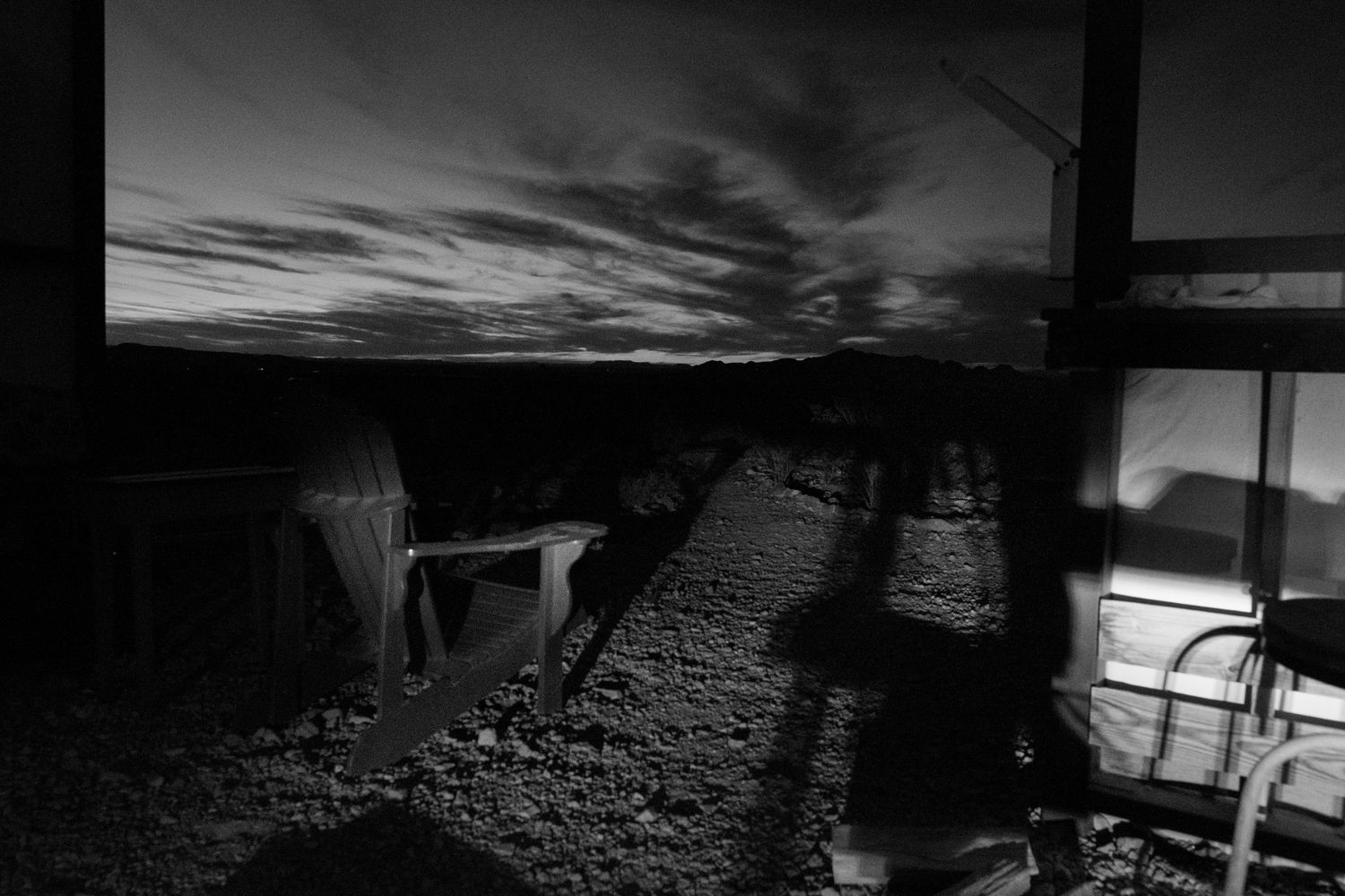 Black and white twilight photo of a campsite with tents and equipment casting long shadows under a cloudy sky.