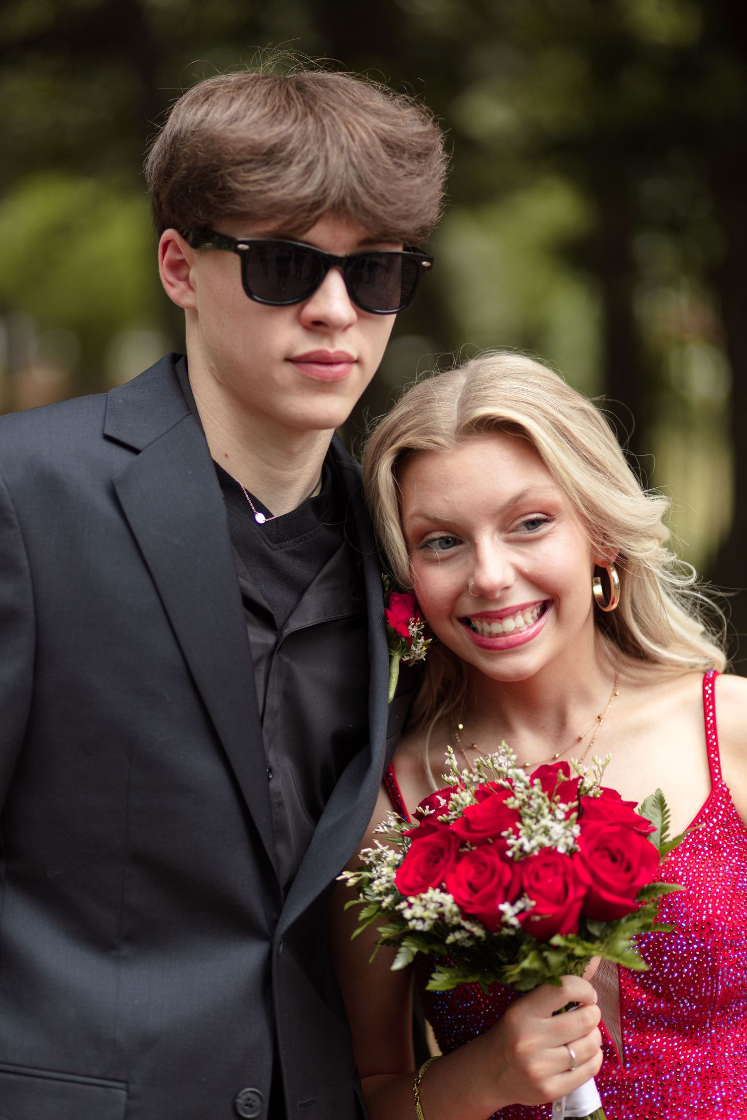 Smiling prom couple, boy in black suit with sunglasses and girl in red dress holding rose bouquet.