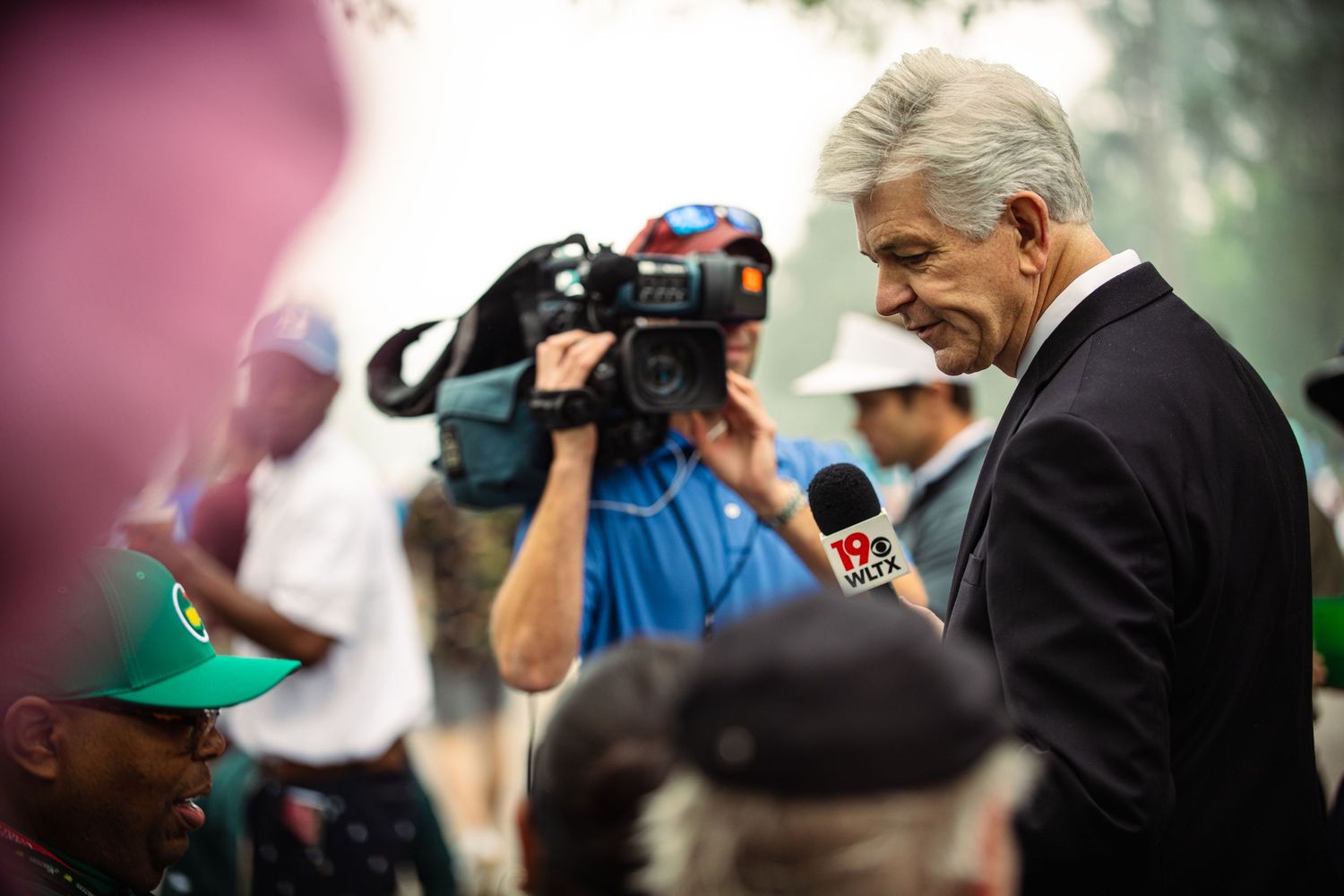 A politician in a suit speaks into a microphone at an outdoor press event with a TV camera crew nearby.