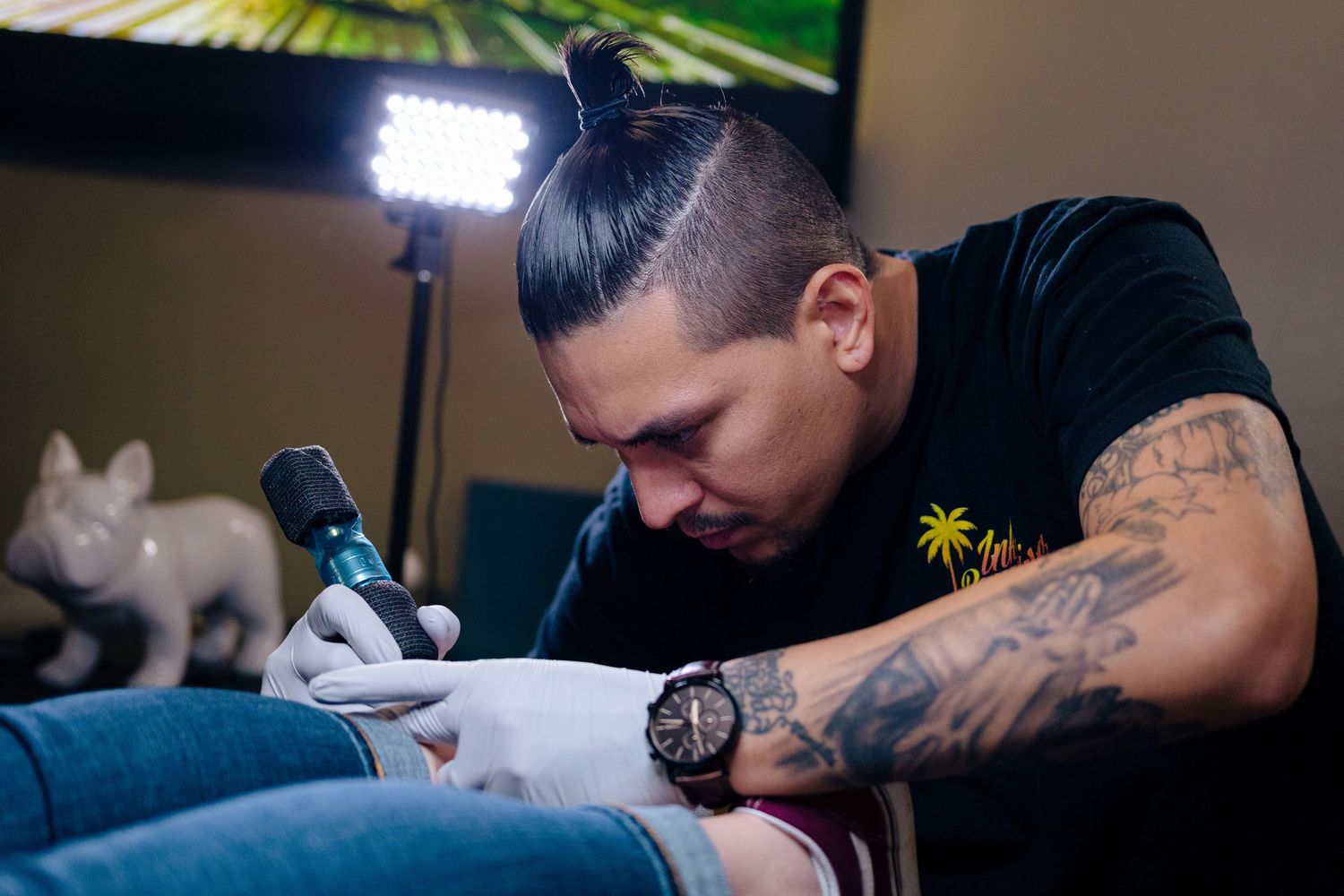 A tattooed male artist carefully applies a tattoo using a tattoo machine while wearing gloves in a professional studio.