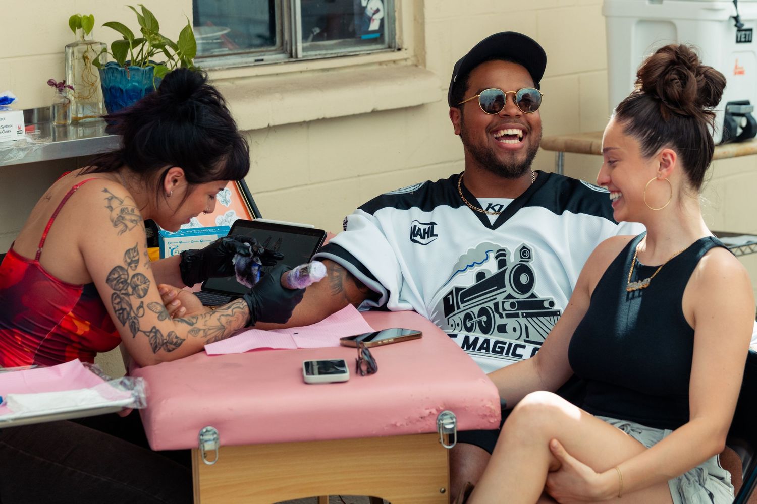 Man laughing joyfully while receiving a tattoo outdoors, accompanied by a smiling woman beside him.