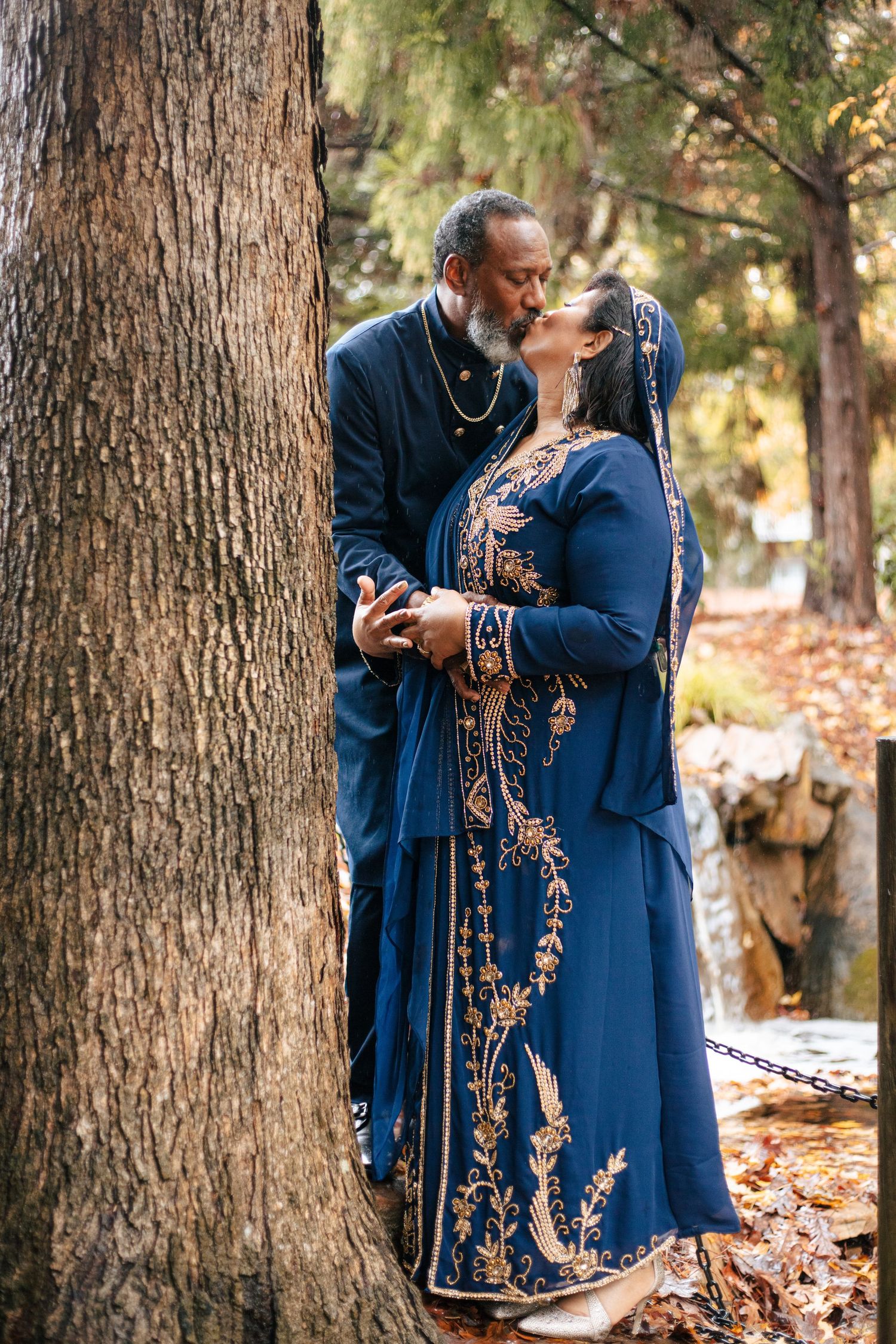Couple in elegant blue traditional attire shares a romantic kiss beside a large tree outdoors.