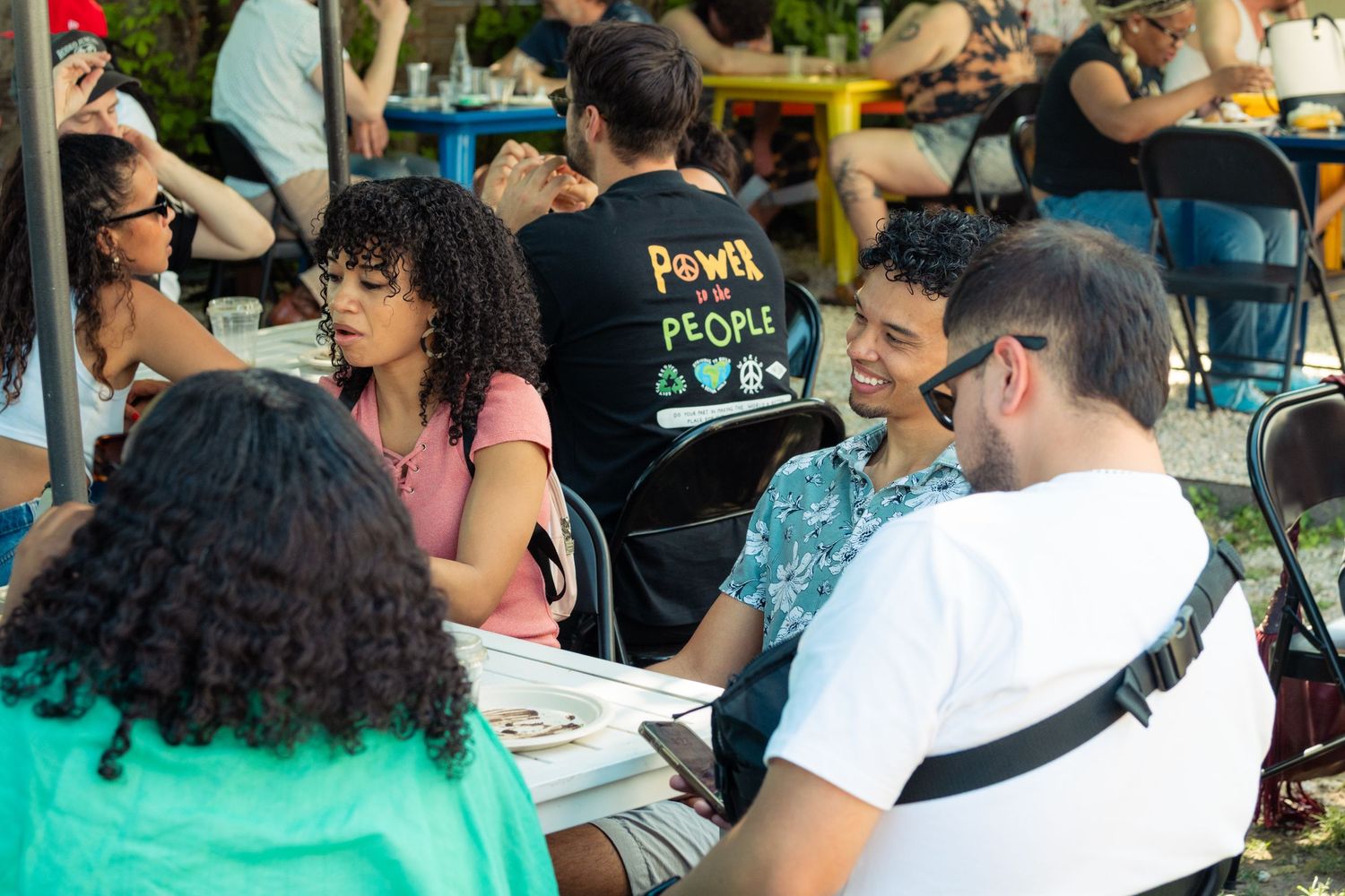 Young adults laughing and talking at an outdoor table, one wearing a Power to the People shirt.