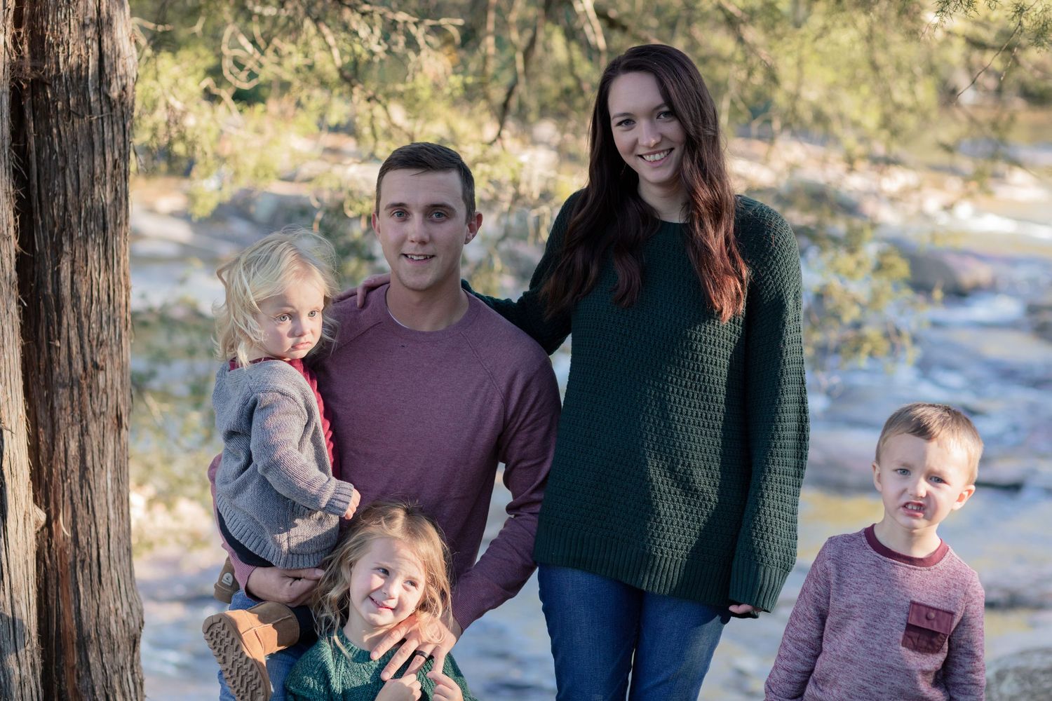 Young family of five posing outdoors near a tree by a rocky riverbank.