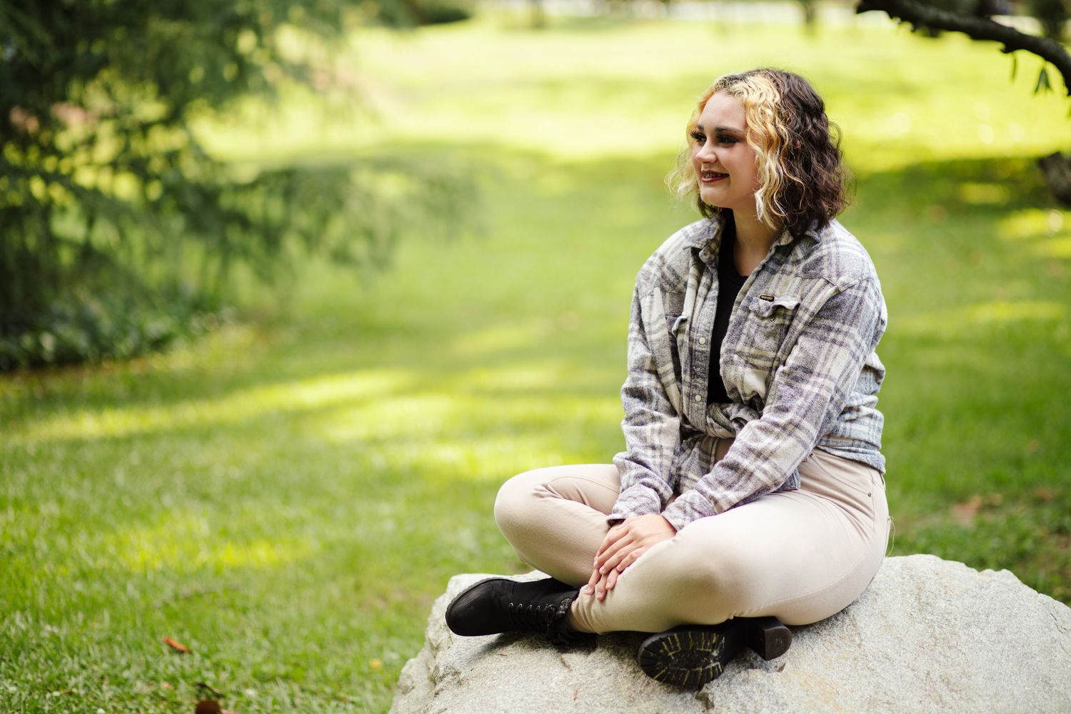 Young blonde woman in plaid shirt sitting cross-legged on rock in park.