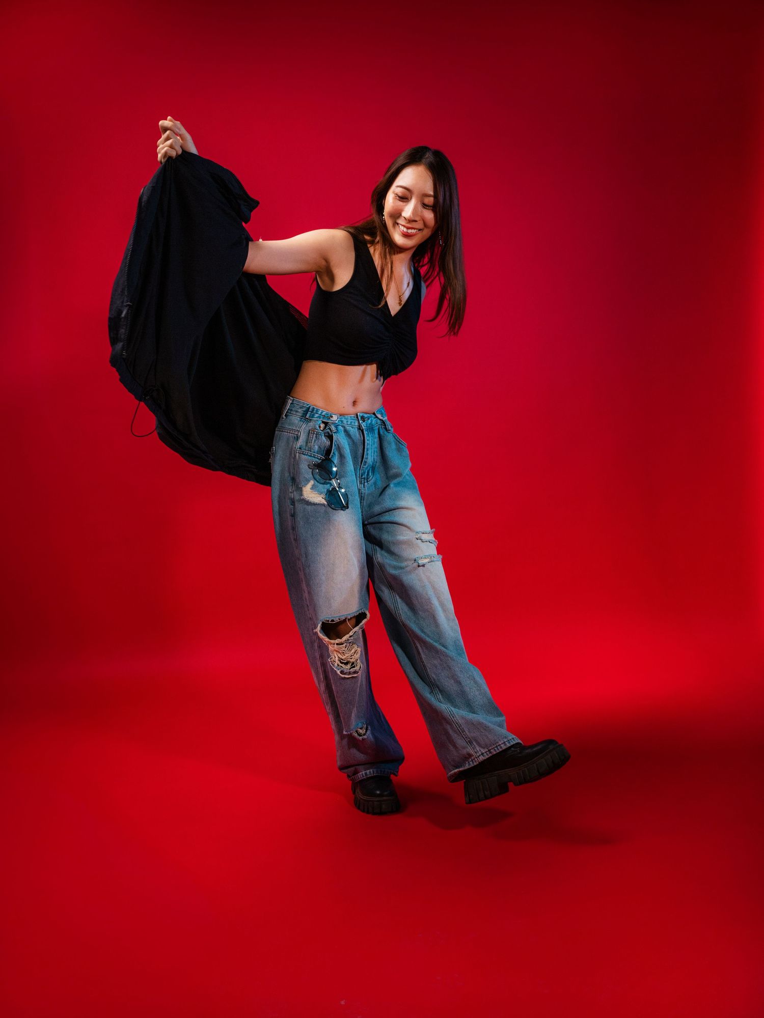 Smiling Asian woman twirls black jacket wearing ripped jeans and crop top against a vivid red backdrop.