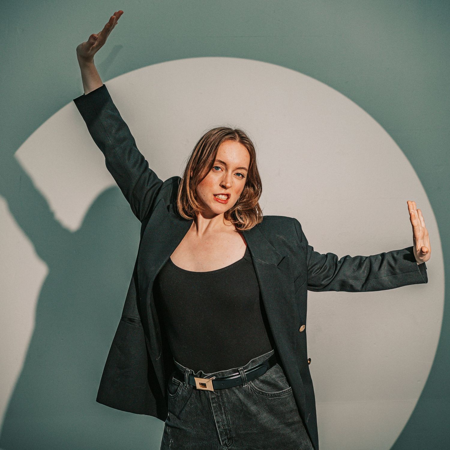 Woman in dark blazer posing with arms raised in front of circular white backdrop.