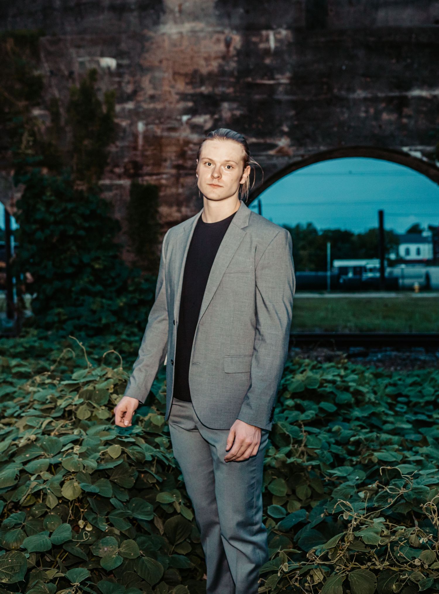 Young man in a grey suit stands among green ivy beneath a brick archway at dusk.