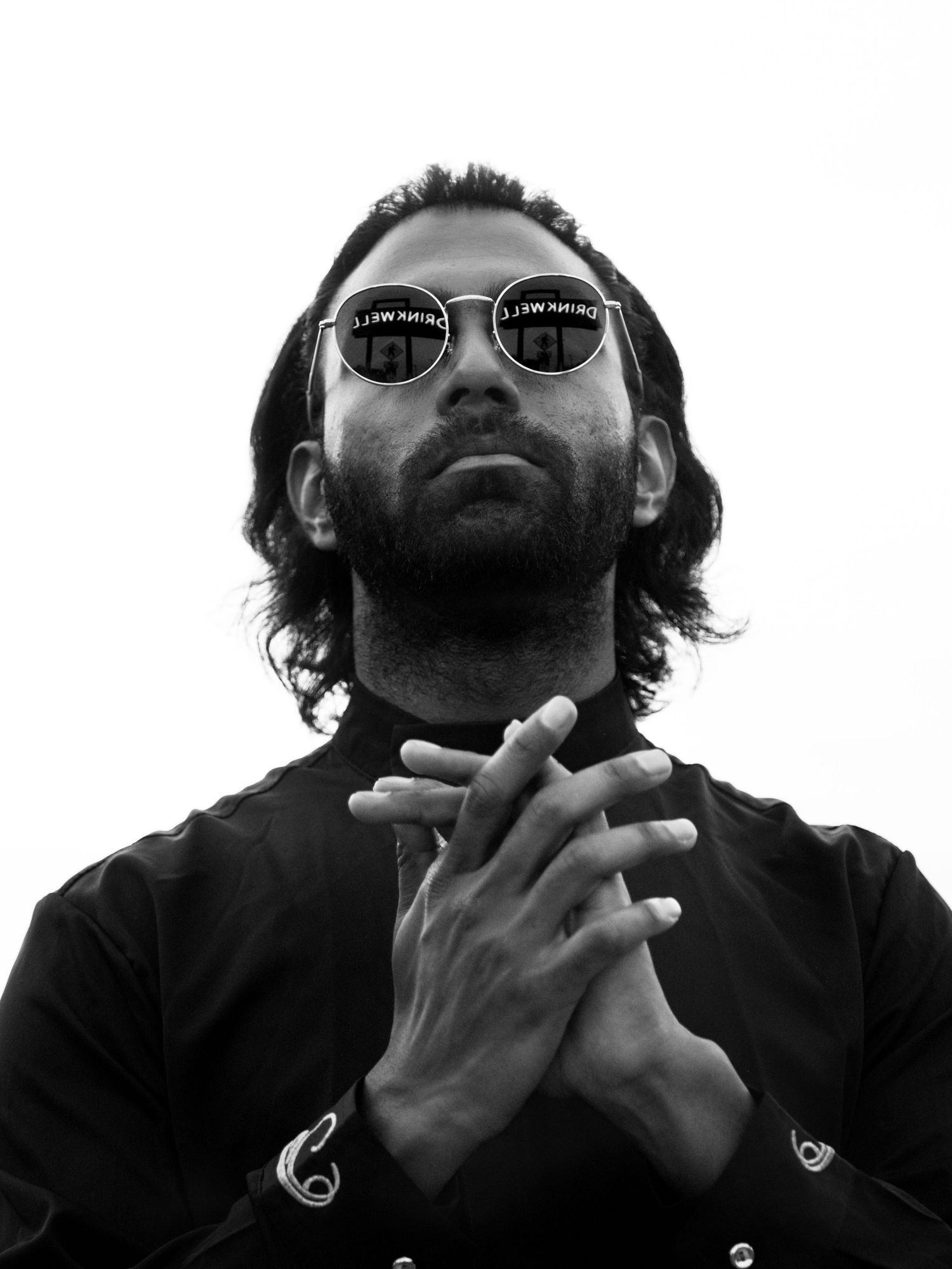 Black-and-white studio portrait of a bearded man in sunglasses with hands clasped, looking upward.