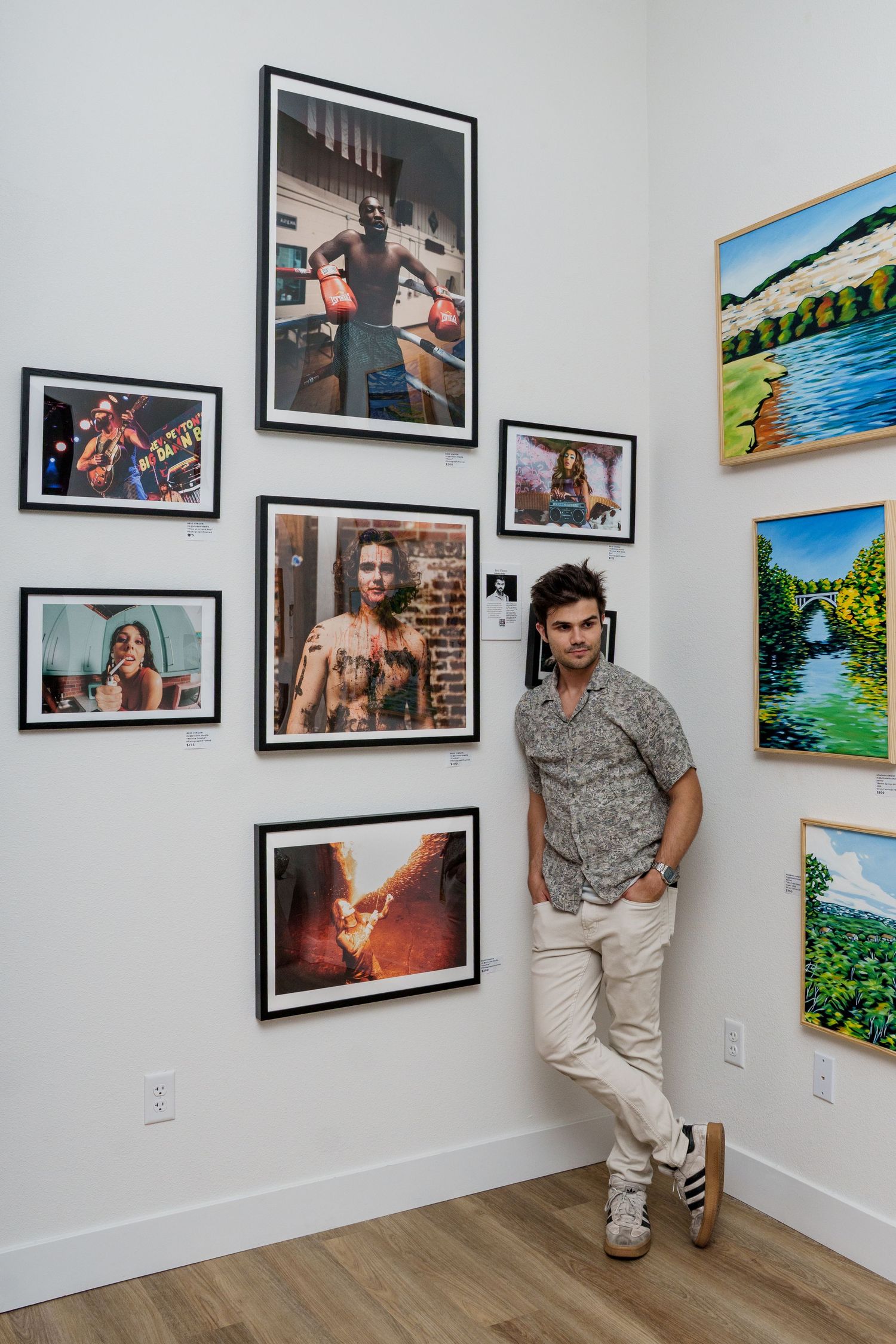 Man standing in art gallery beside framed photography prints displayed on white wall with colorful landscape paintings nearby.