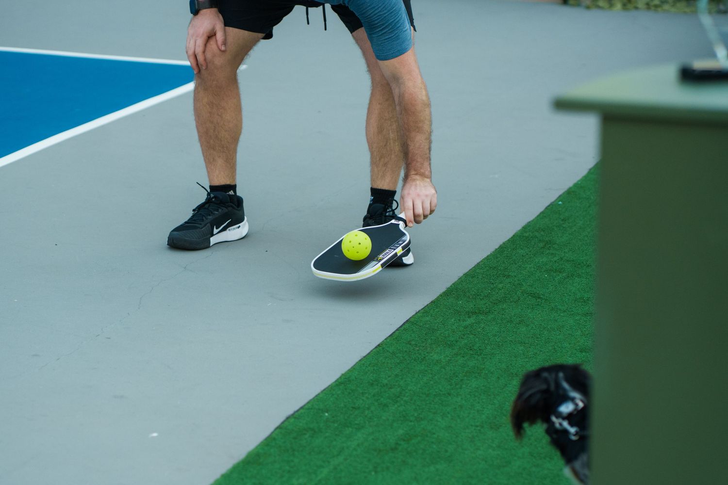 A tennis player in Nike shoes picks up a yellow tennis ball with their foot on a gray court surface.