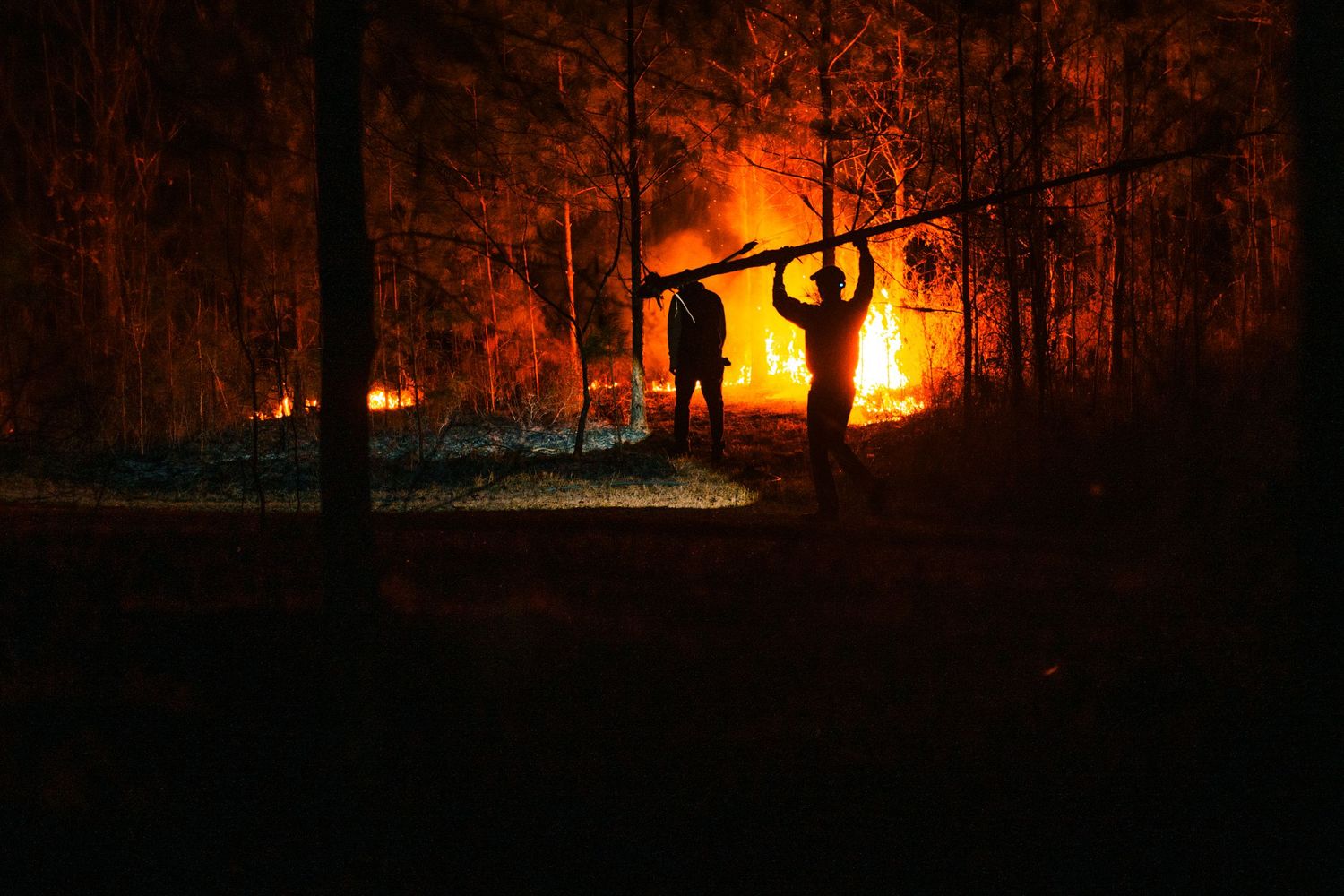 Two silhouetted figures carry a log near a large forest fire burning at night.