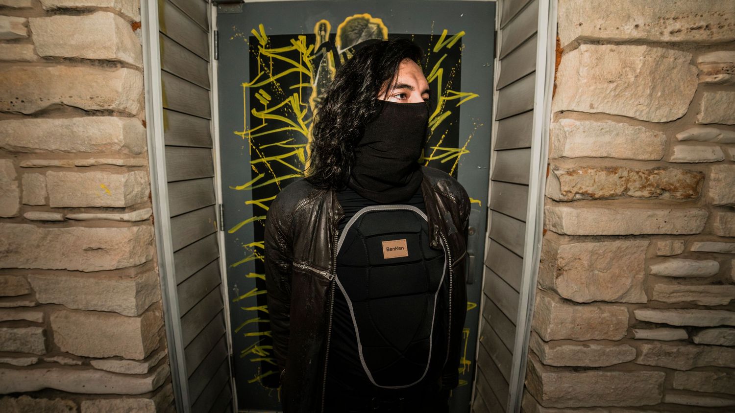 Person in black mask and jacket wrapped in yellow caution tape in doorway.