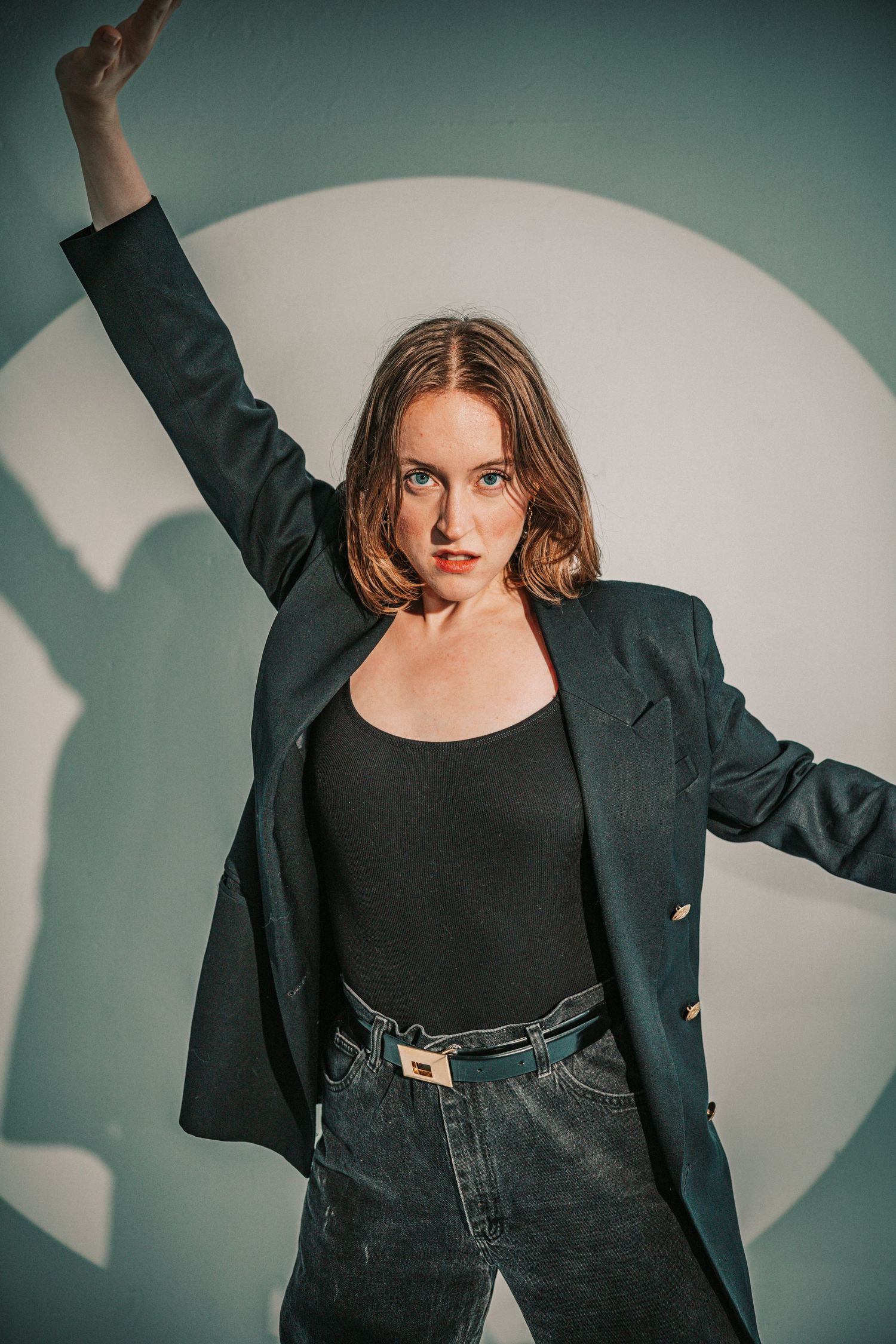 Young woman in dark blazer and jeans poses dynamically with arm raised against light background.