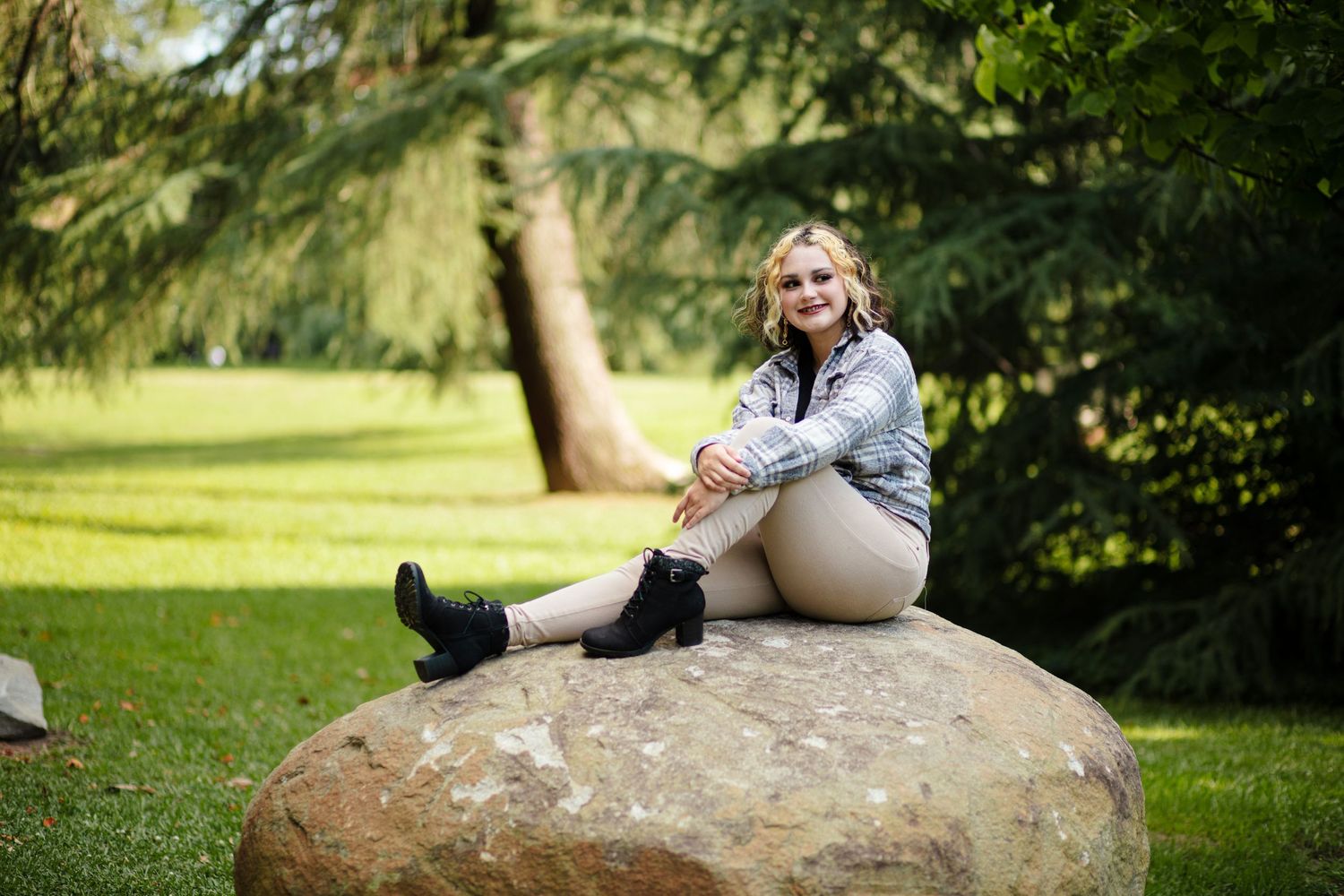 Blonde teen girl in plaid jacket posing on large boulder in sunny park.